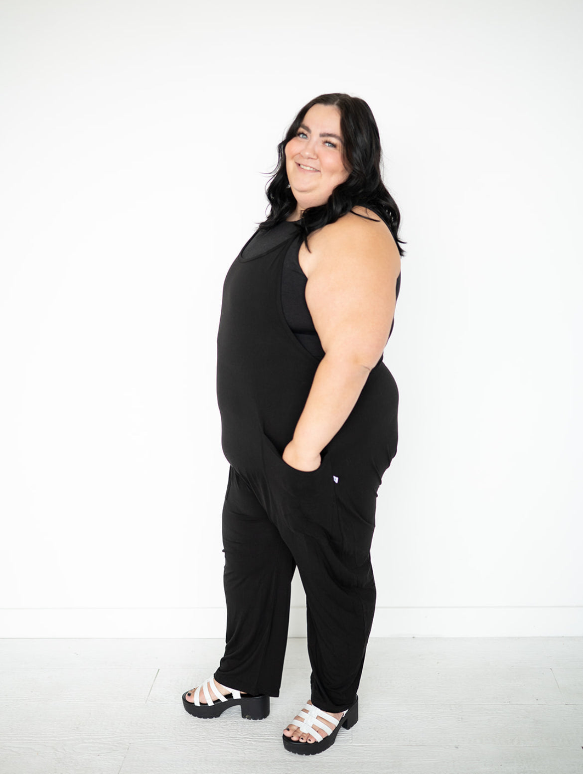 Plus-size woman in black sleeveless overalls and white platform sandals.