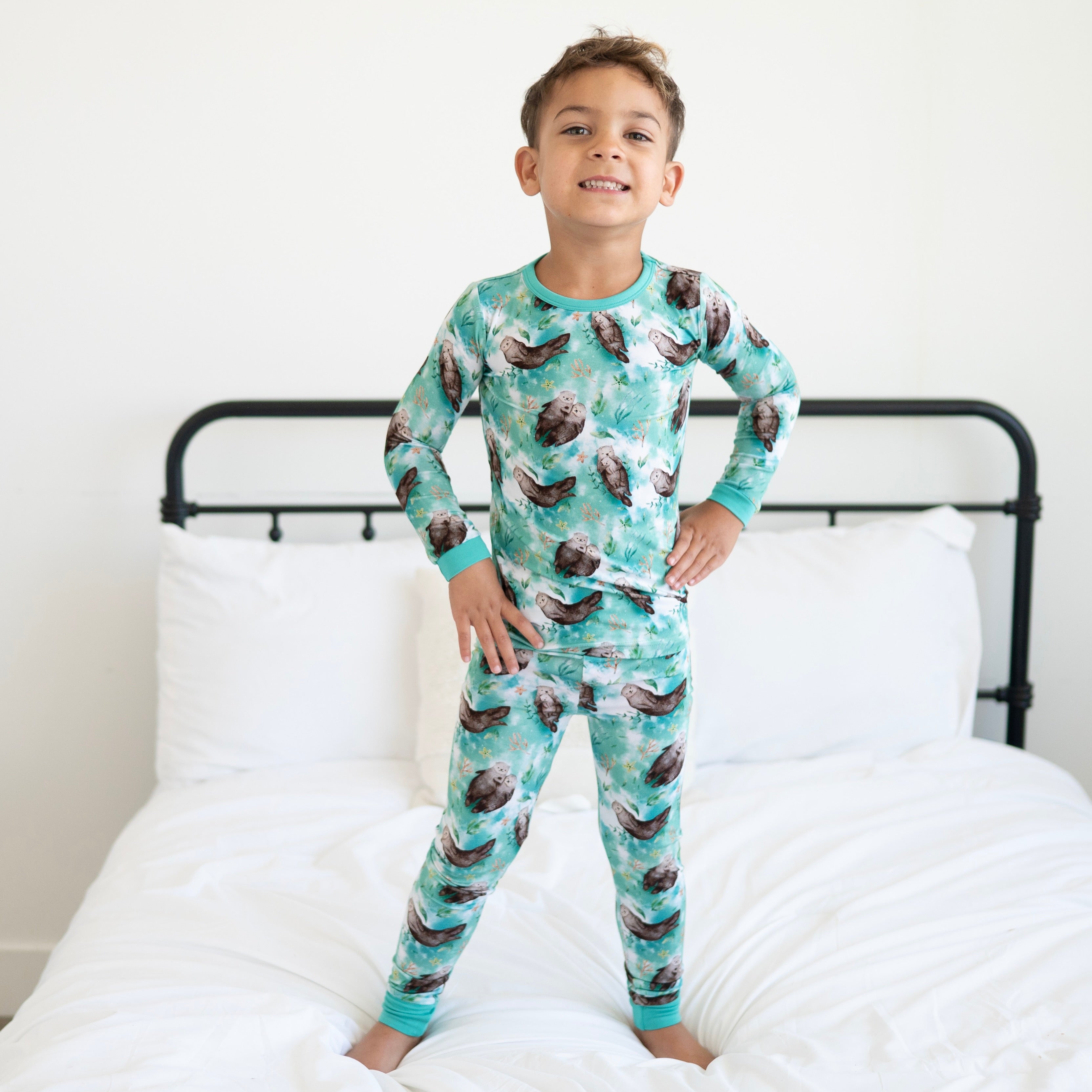 A young boy stands on a bed wearing a long-sleeved pajama set with an otter print.