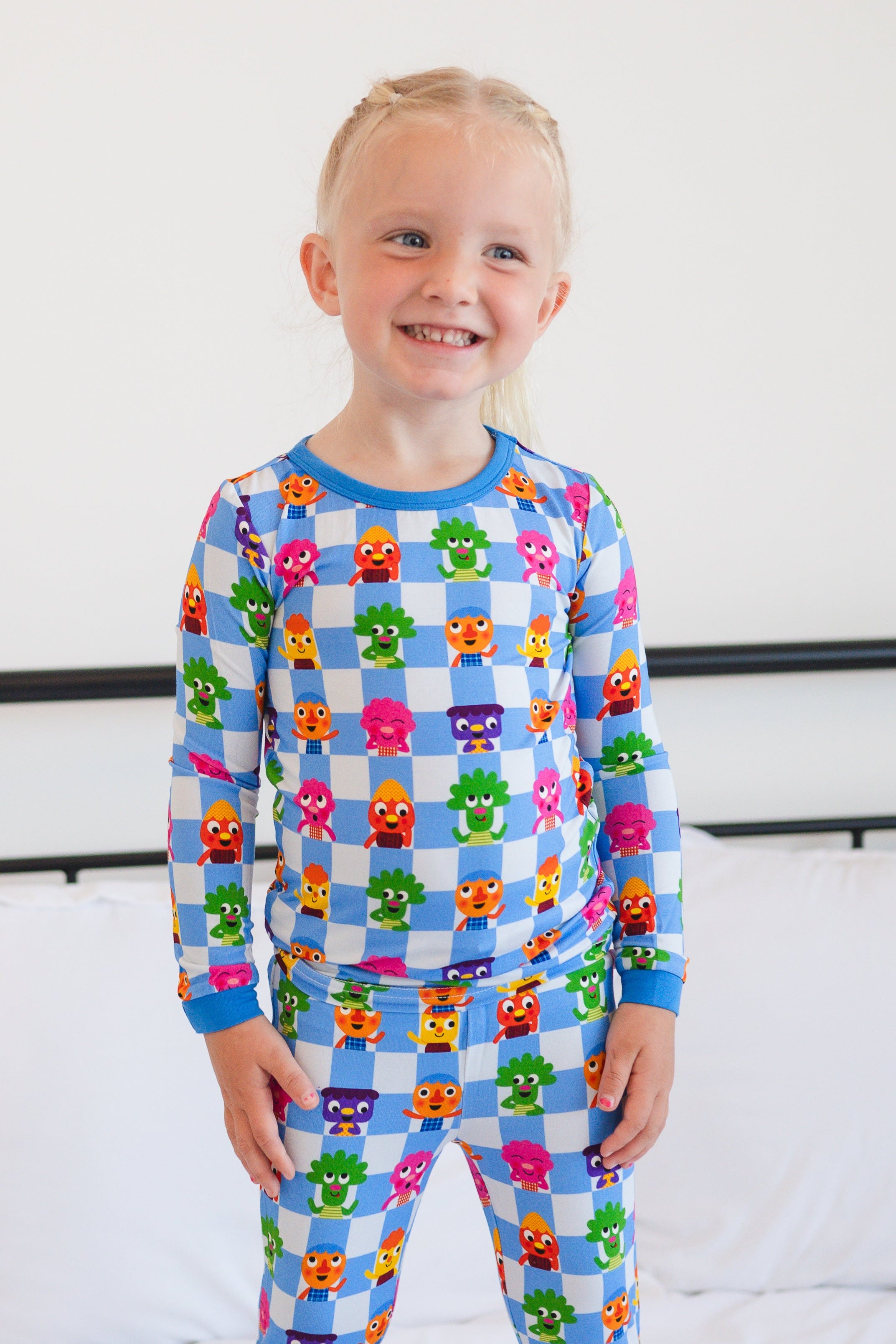 A young girl stands on a bed wearing blue pajamas with a checkered pattern and cartoon characters.
