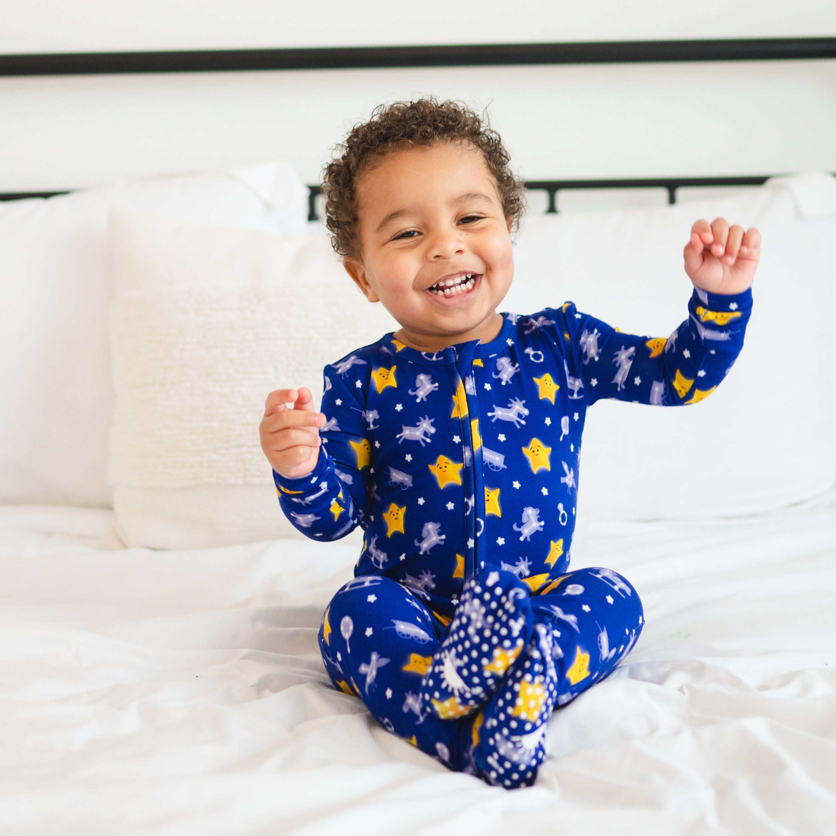 A smiling baby boy sits on a white bed wearing blue pajamas with stars and unicorns.