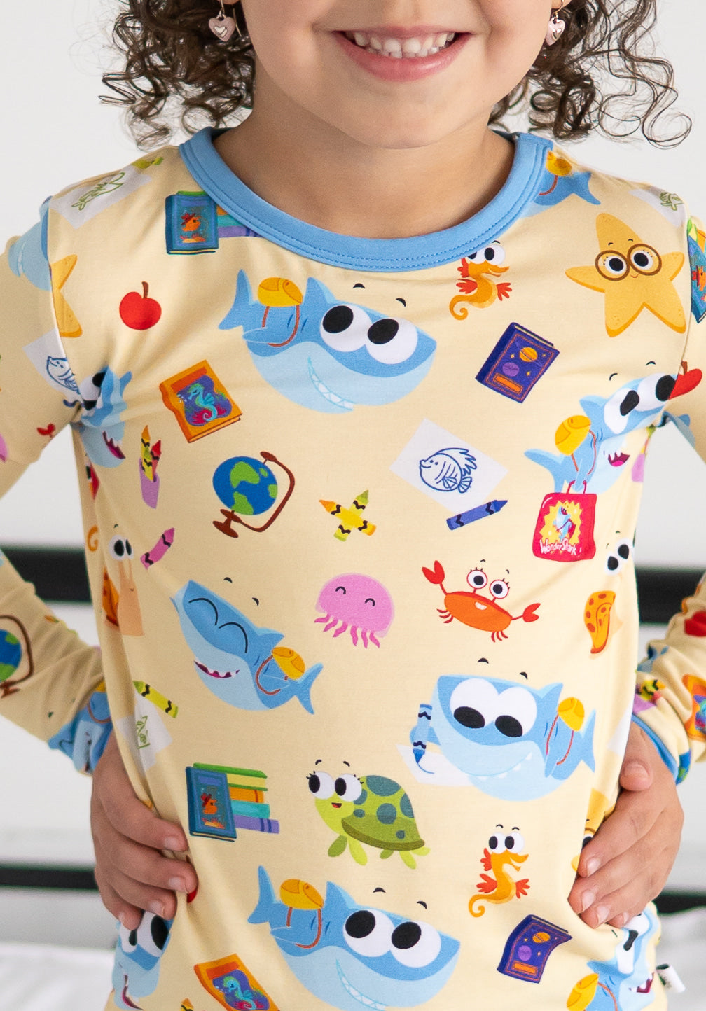 A child wears a yellow long-sleeved pajama top with a print of cartoon sharks, sea creatures, and school supplies.