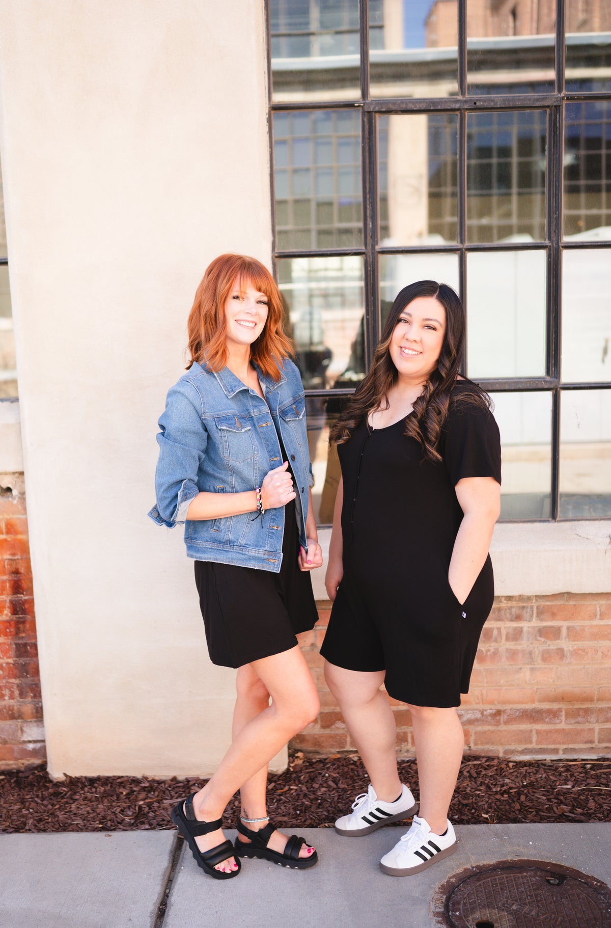 Two women stand in front of a building, one in a denim jacket and black romper, the other in a black romper.