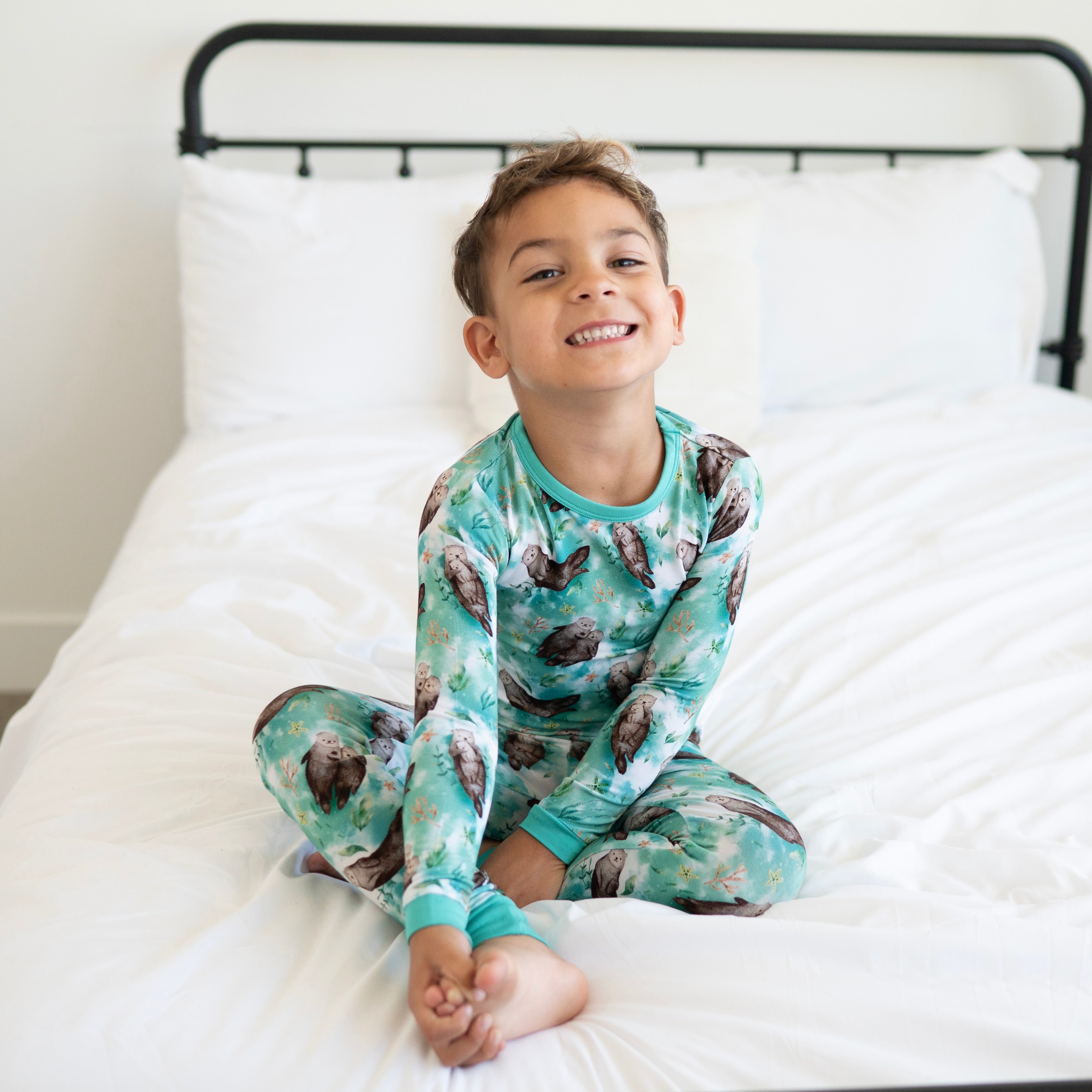 A young boy smiles while sitting on a bed wearing a teal pajama set with an otter print.