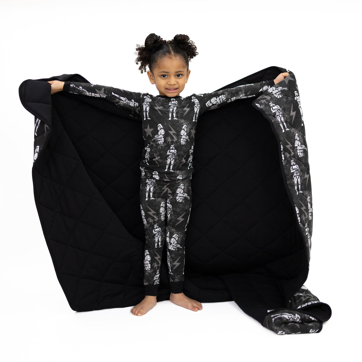 A child in Stormtrooper pajamas holds a black quilted blanket with a Stormtrooper print.