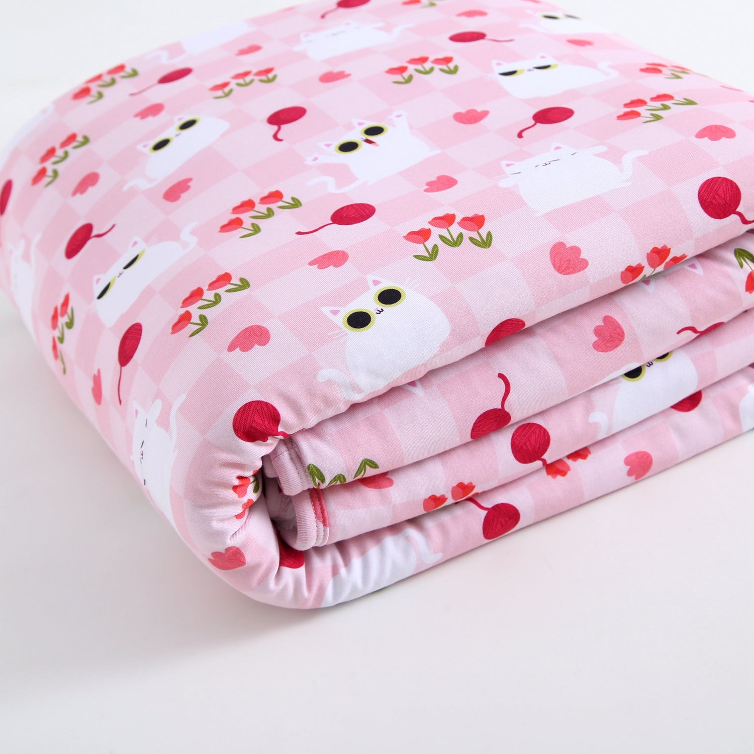A folded pink blanket with a checkered pattern featuring white cats and red yarn balls.