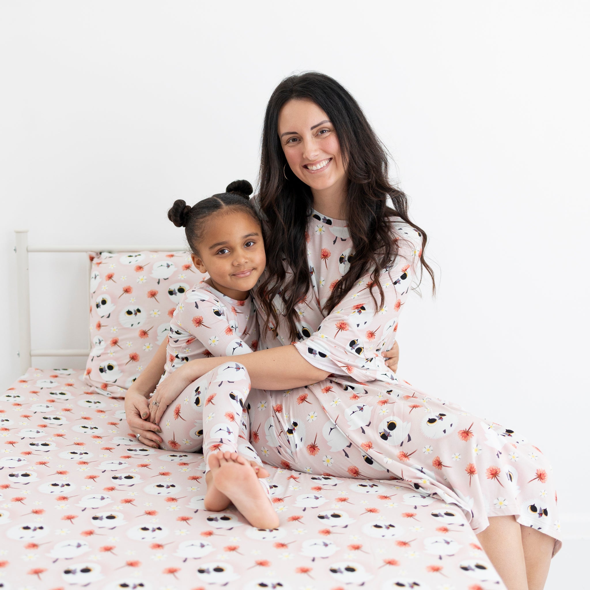 A woman and child in matching pink Puffin Rock pajamas sit on a bed with a matching pillow.