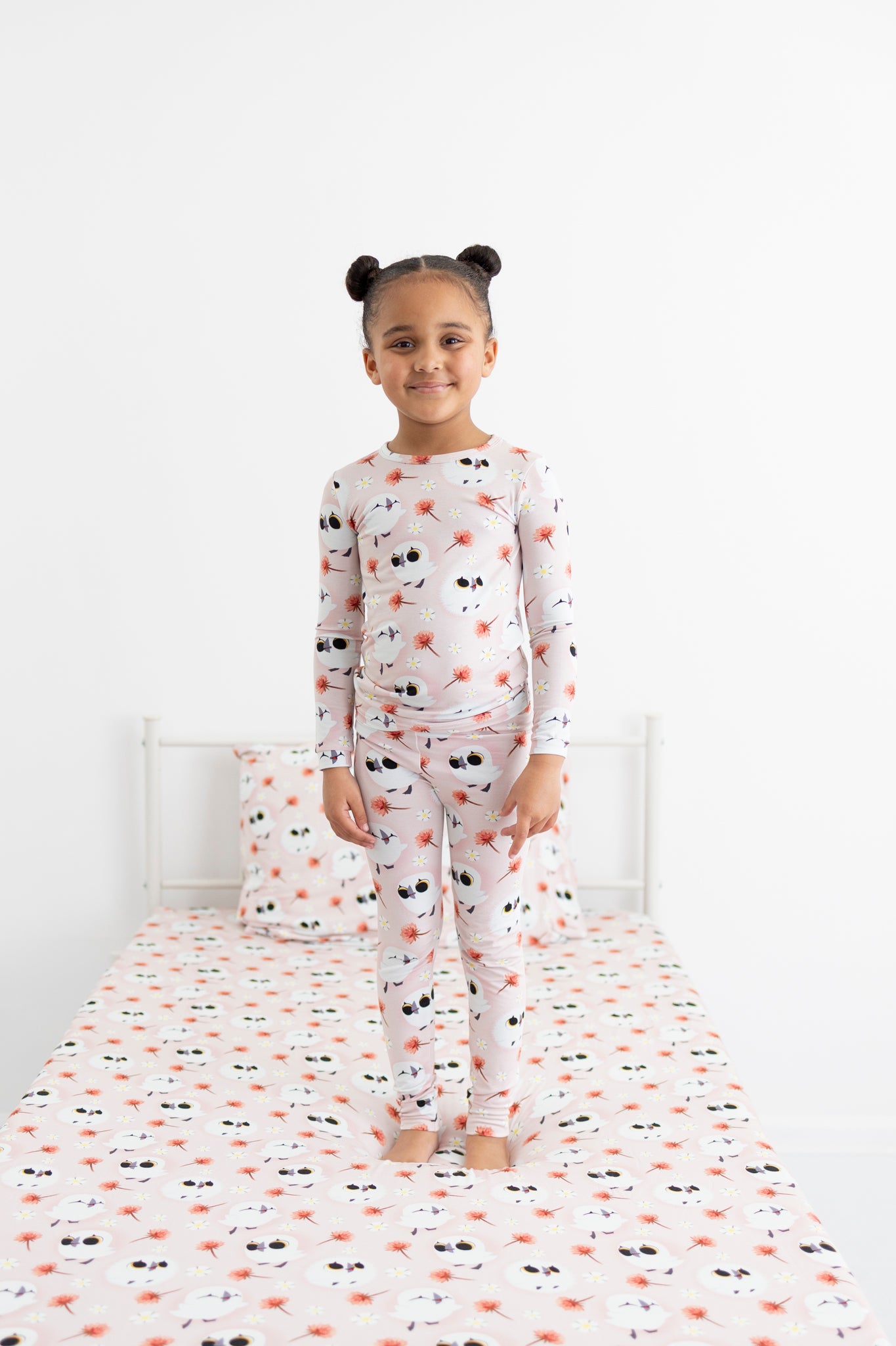 A young girl stands on a bed wearing a pink pajama set with a puffin print.