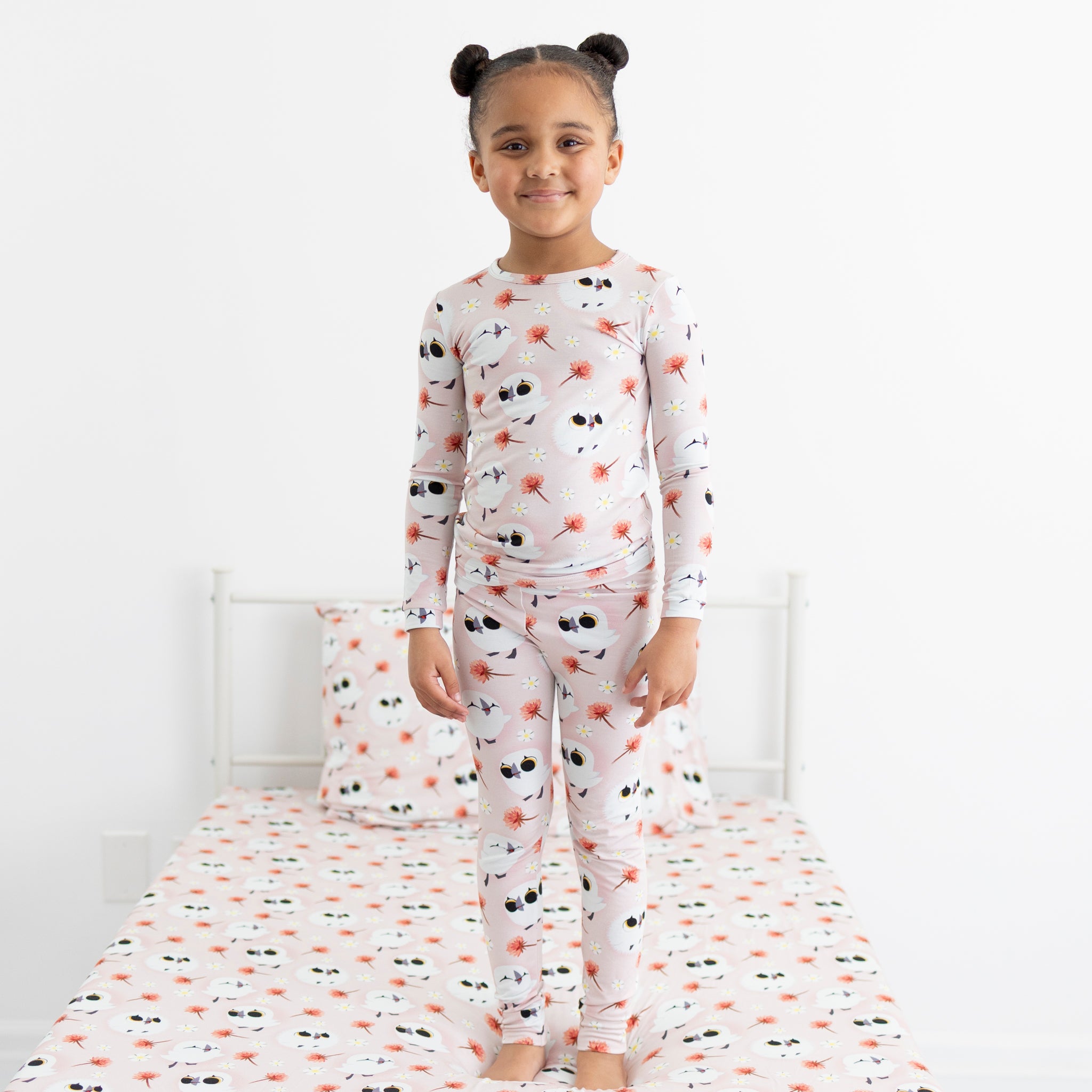 A young girl stands on a bed wearing pink pajamas with a Puffin Rock pattern.