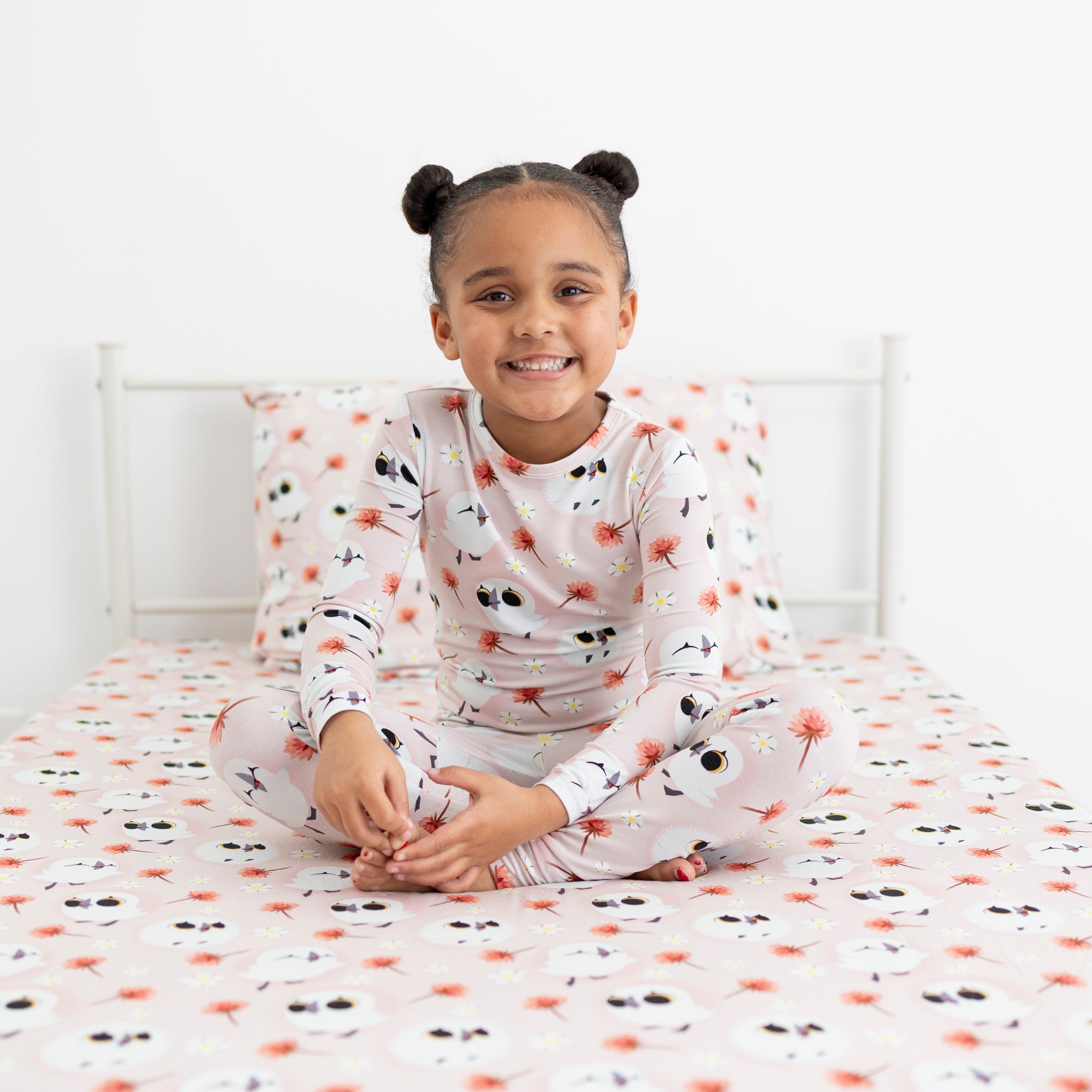 A young girl smiles while sitting cross-legged on a bed in Puffin Rock™ pajamas.