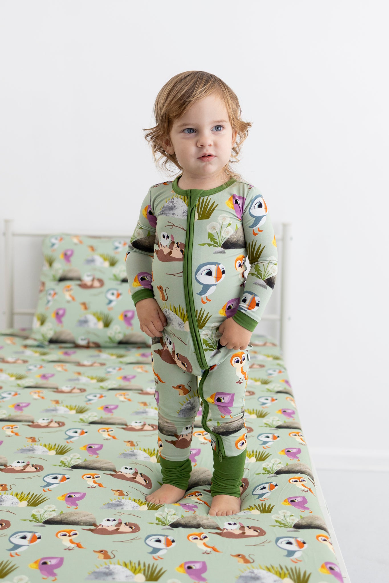 A toddler stands on a bed wearing a green pajama set with a Puffin Rock print.