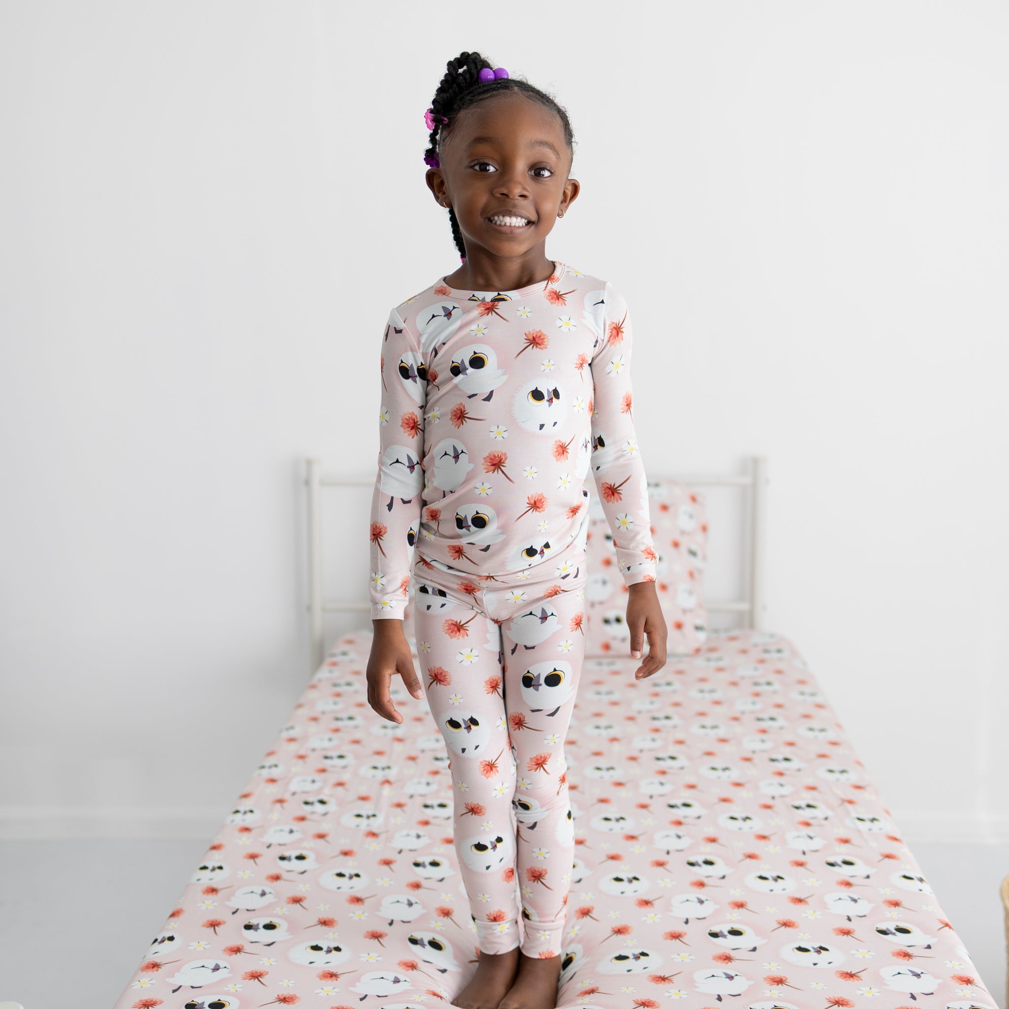 A young girl stands on a bed wearing pink pajamas with a puffin print.