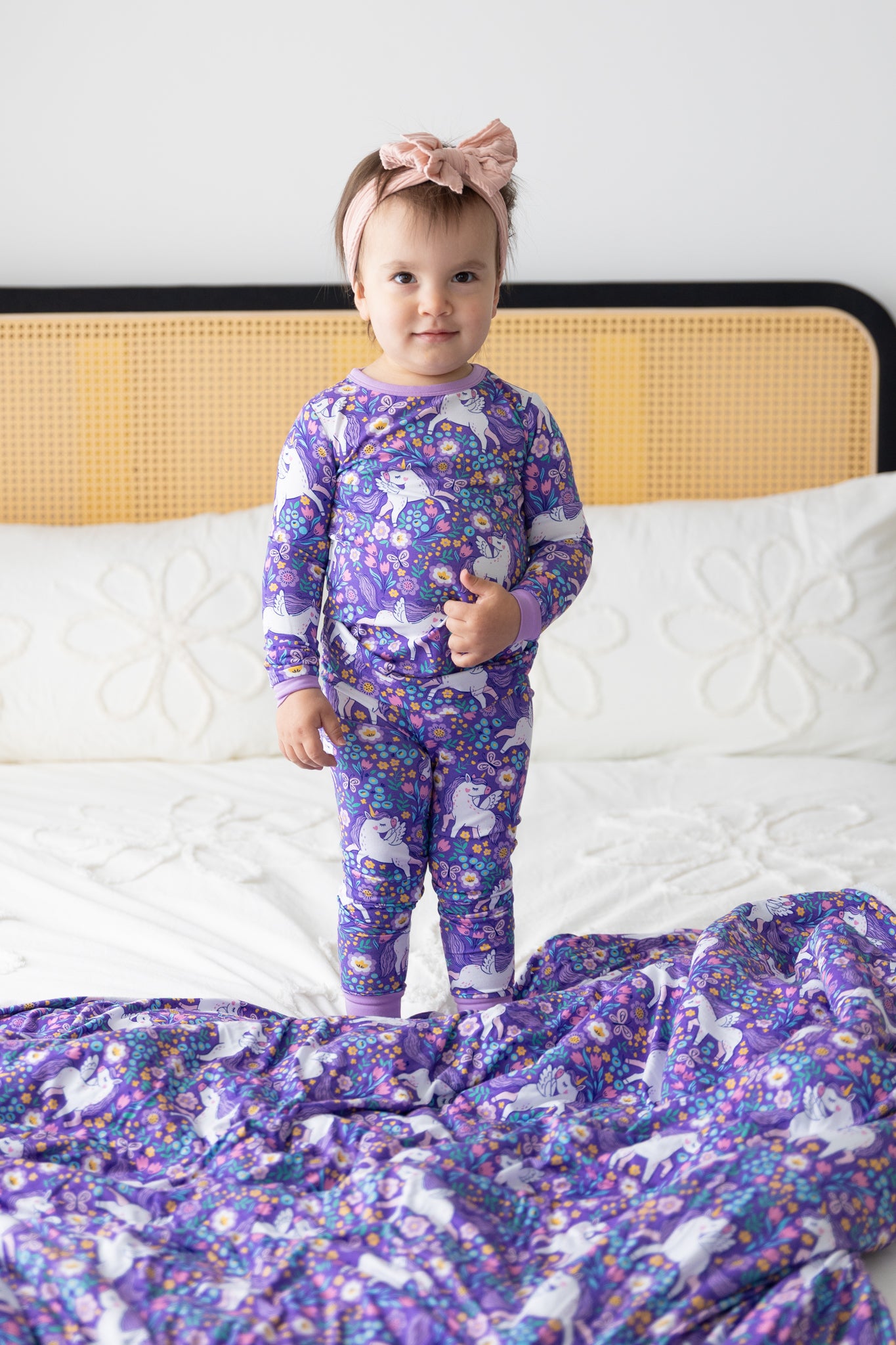 A toddler stands on a bed wearing a purple long-sleeve pajama set featuring a unicorn floral print.