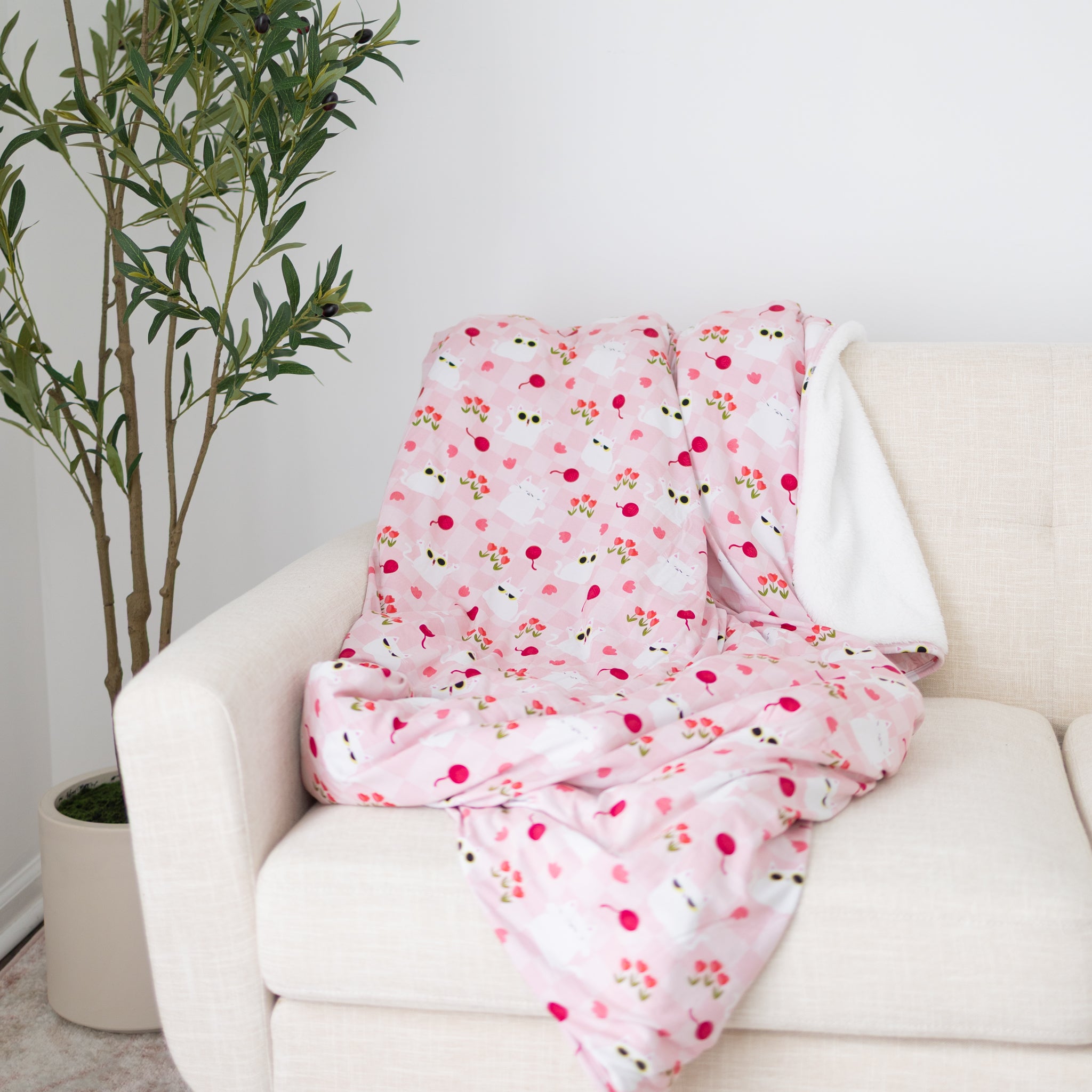 A pink and white checkered blanket with a cat and flower pattern draped over a beige couch.