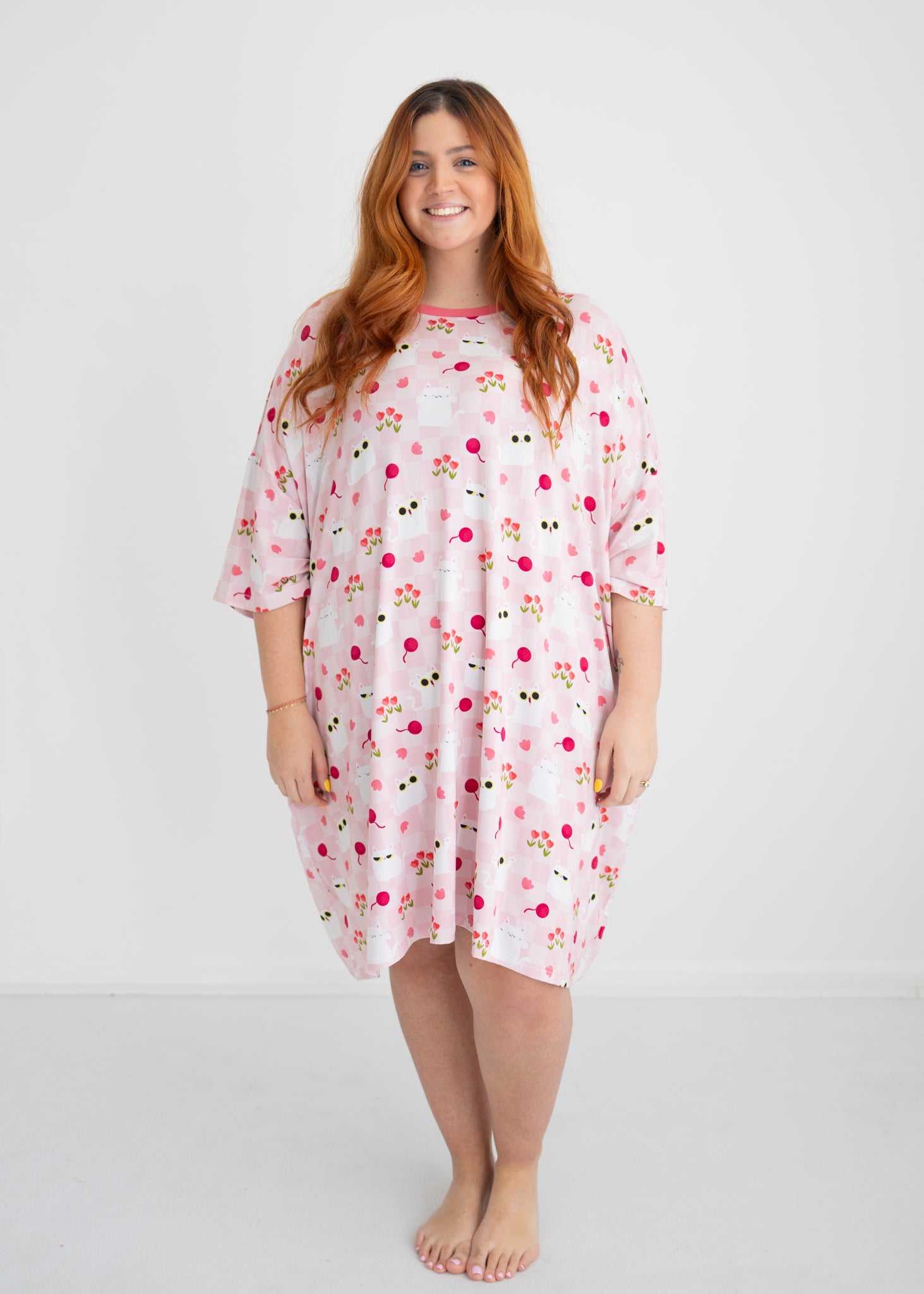 A woman wears an oversized pink sleep tee featuring a repeating pattern of white cats and tulips.