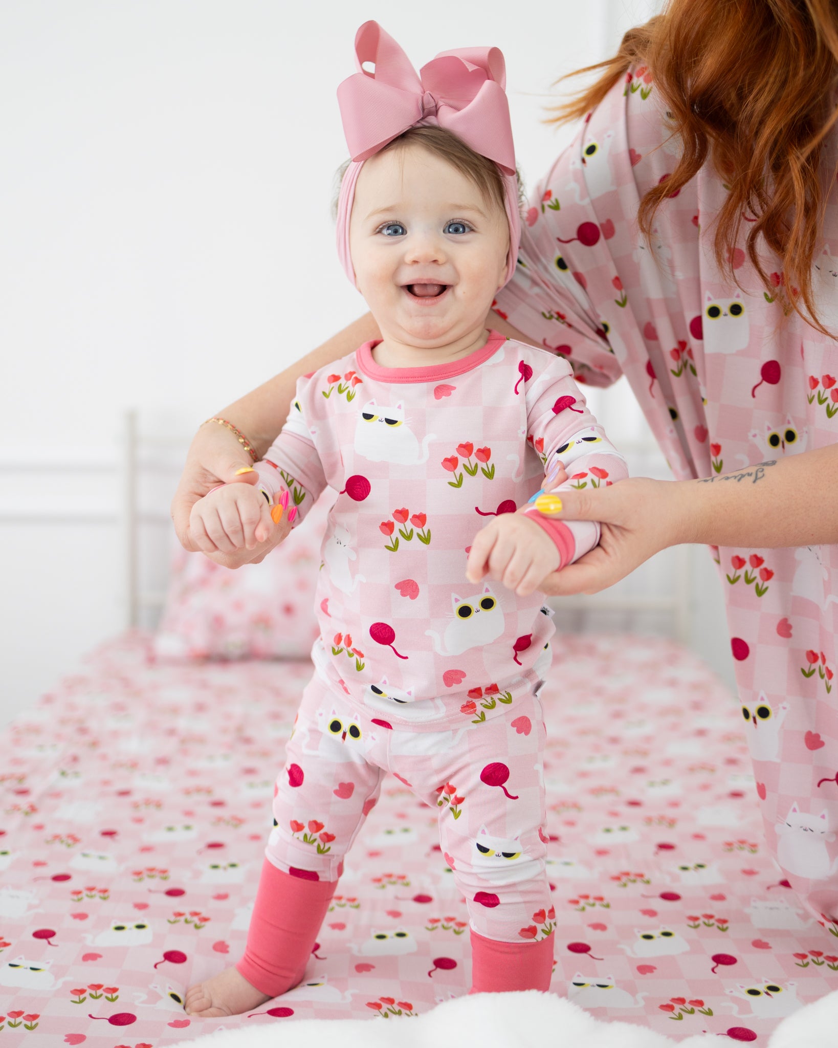A baby wears a pink and white checkered pajama set featuring a cat and tulip print.
