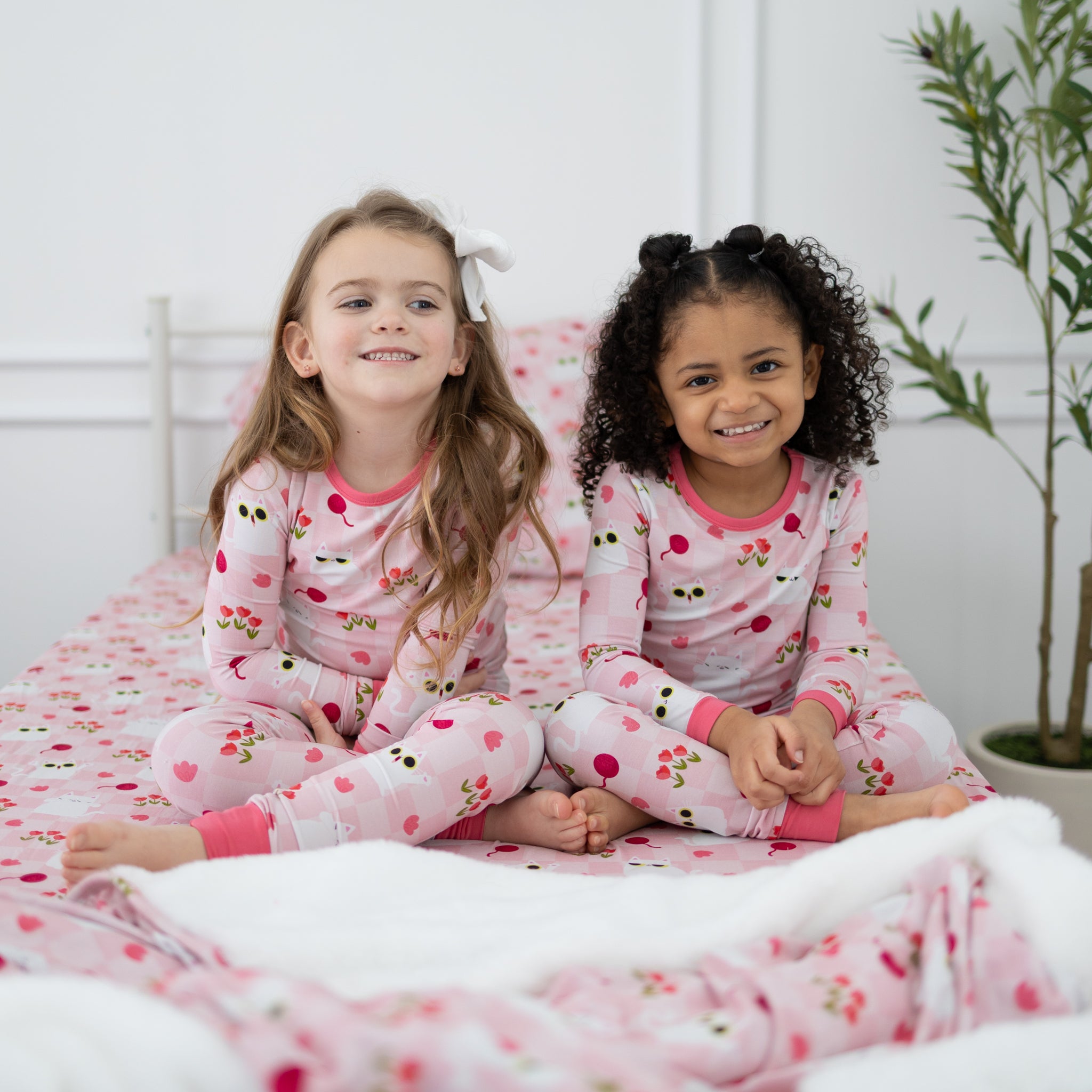 Two young girls sit on a bed wearing matching pink pajamas with a white cat and floral print.