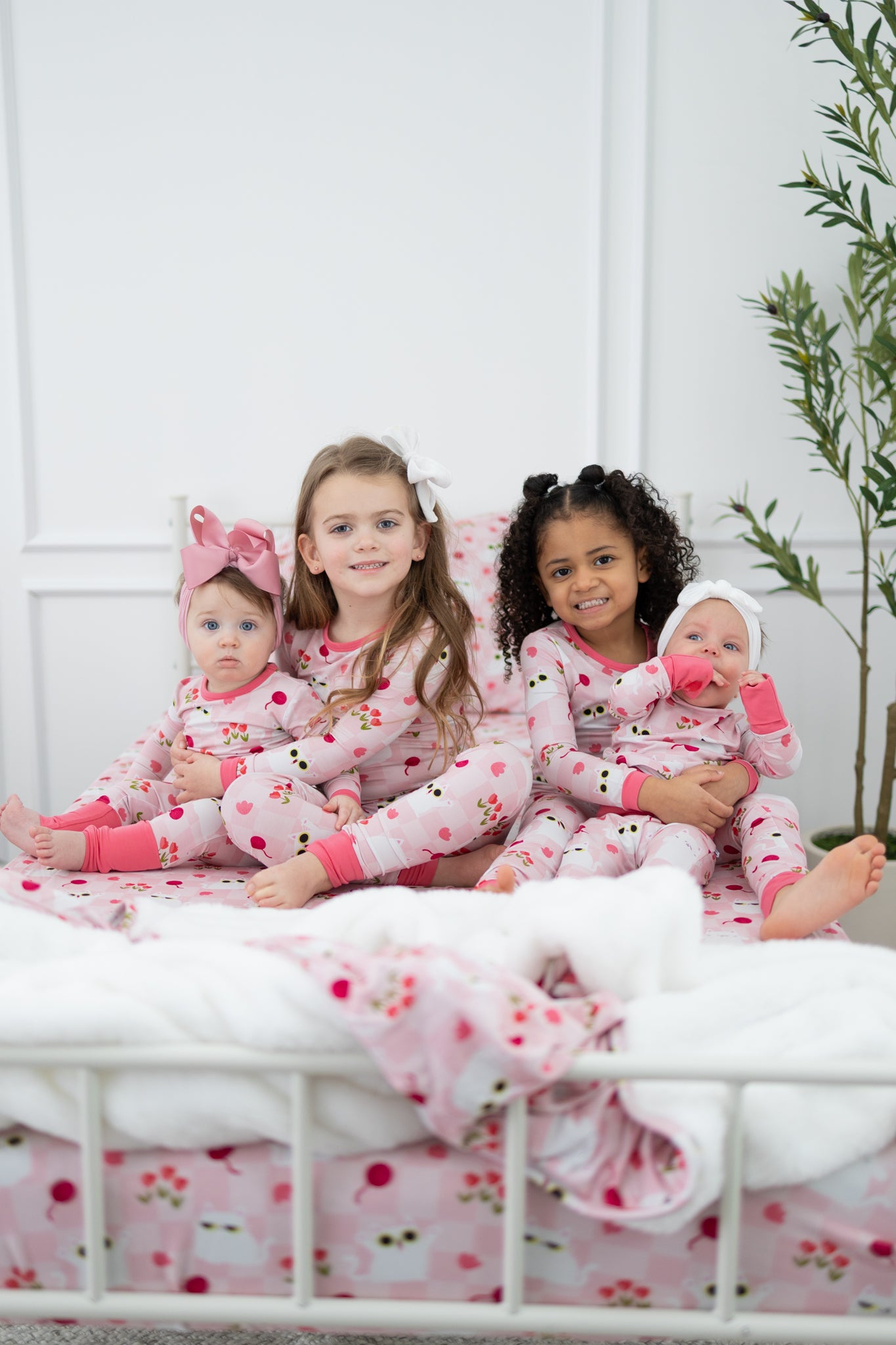 Four children sit on a bed wearing matching pink pajamas with a cat and floral print.