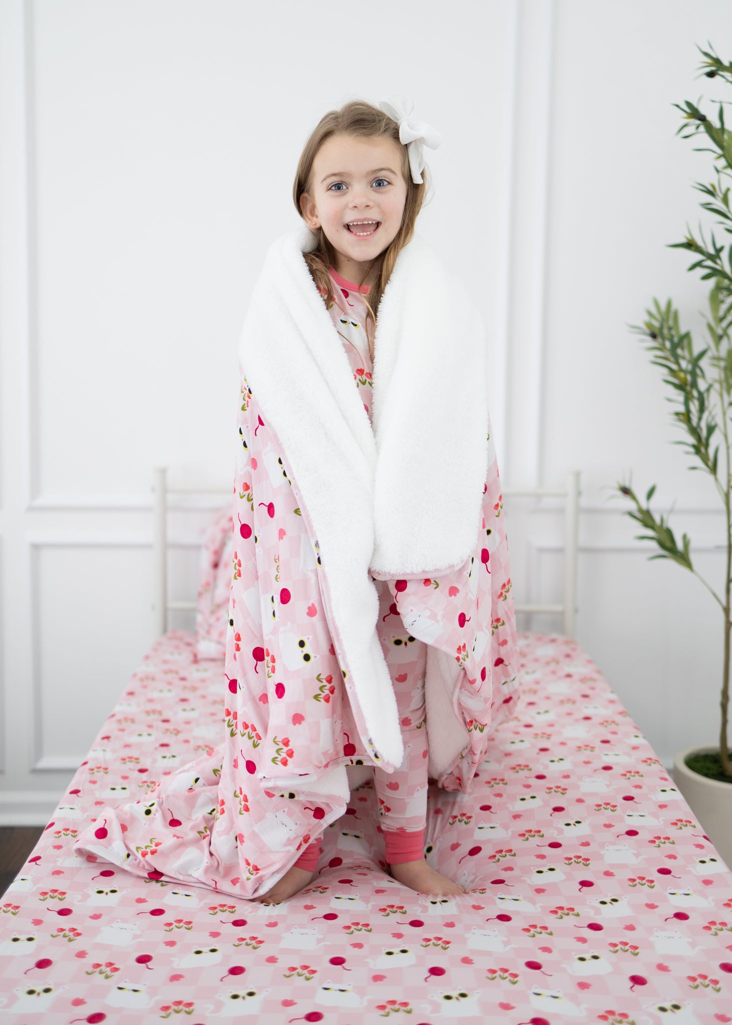 A child stands on a bed wrapped in a pink and white cat-patterned plush blanket.