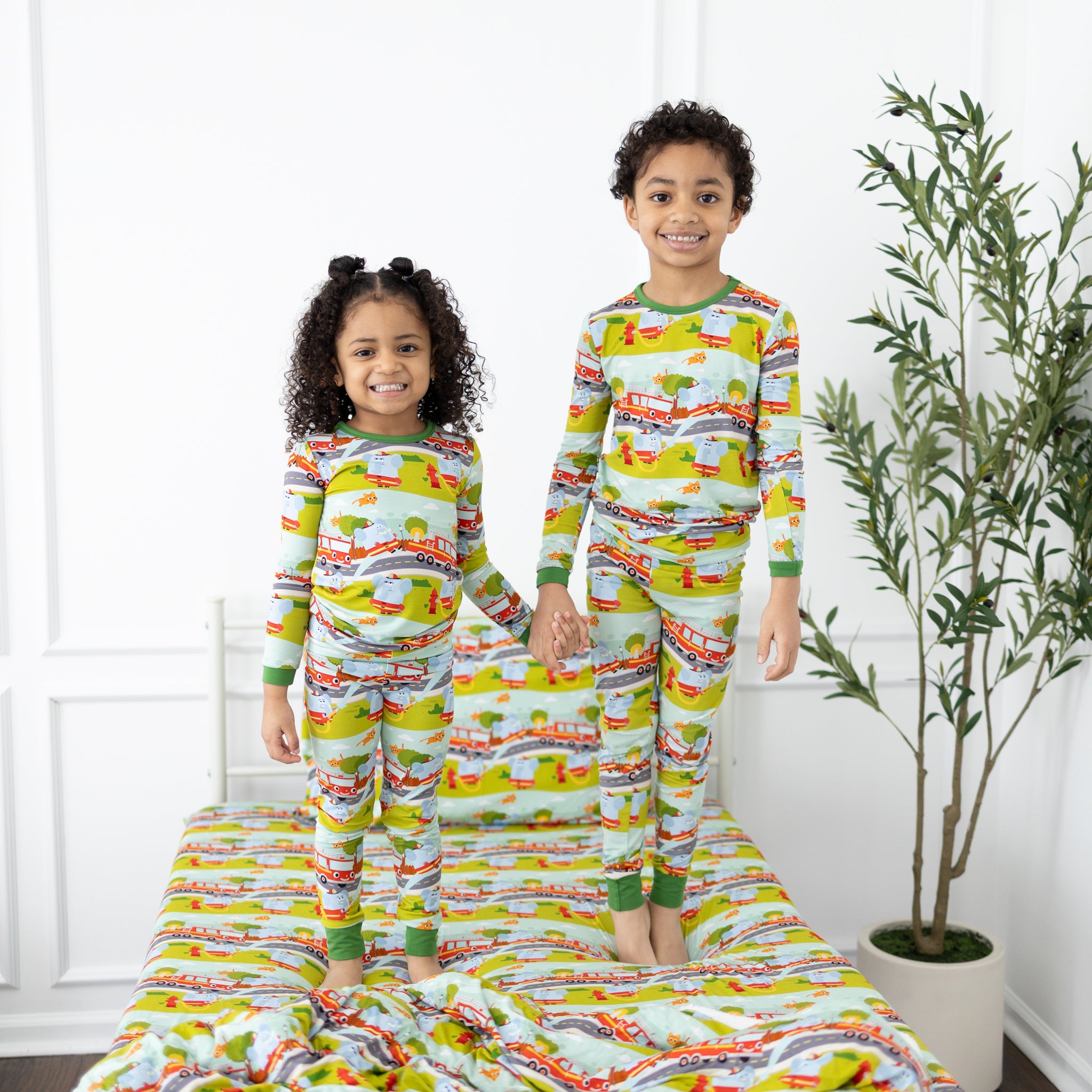 Two children stand on a bed wearing matching long-sleeve pajamas with a firetruck print.