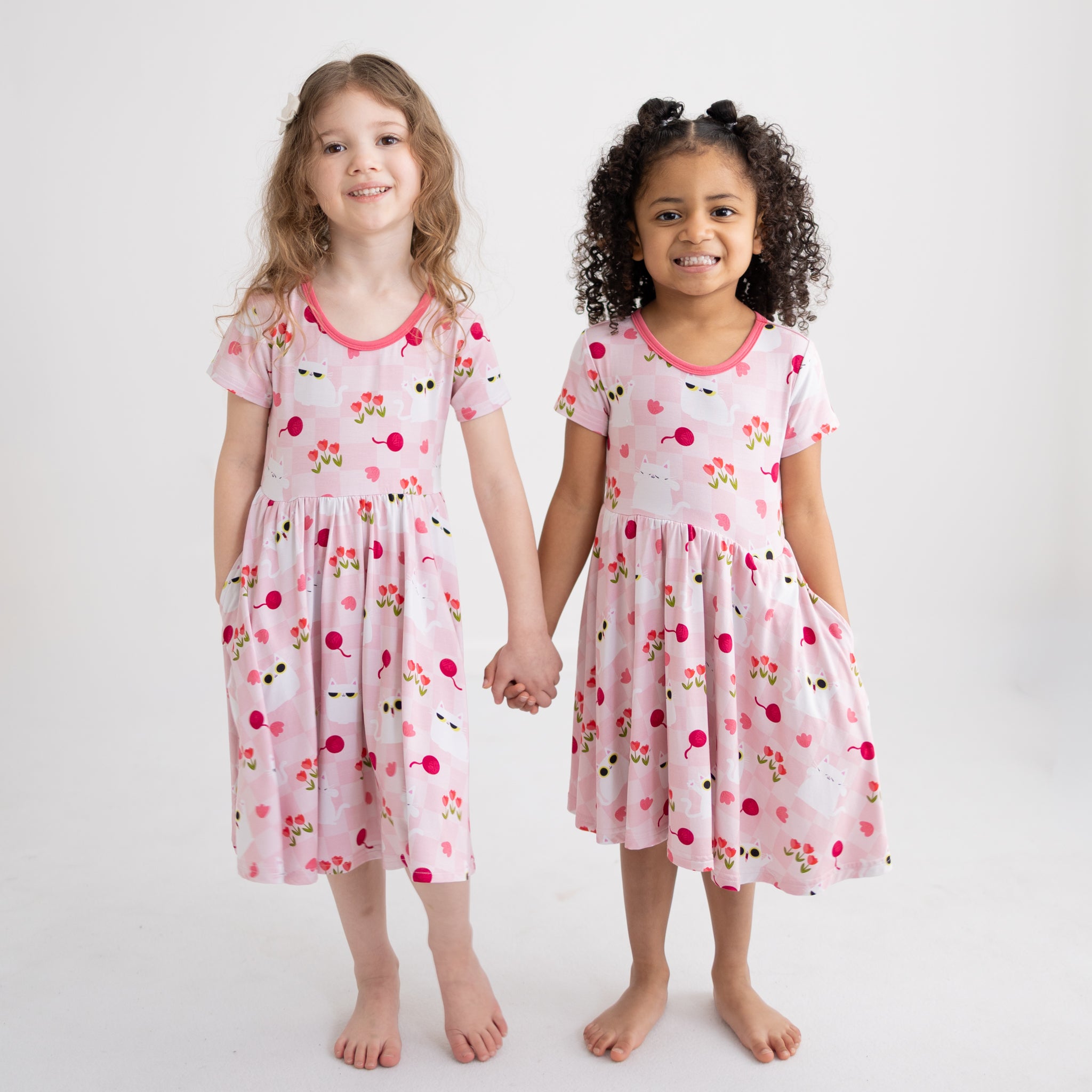 Two girls hold hands wearing pink dresses with a white cat and tulip pattern and side pockets.