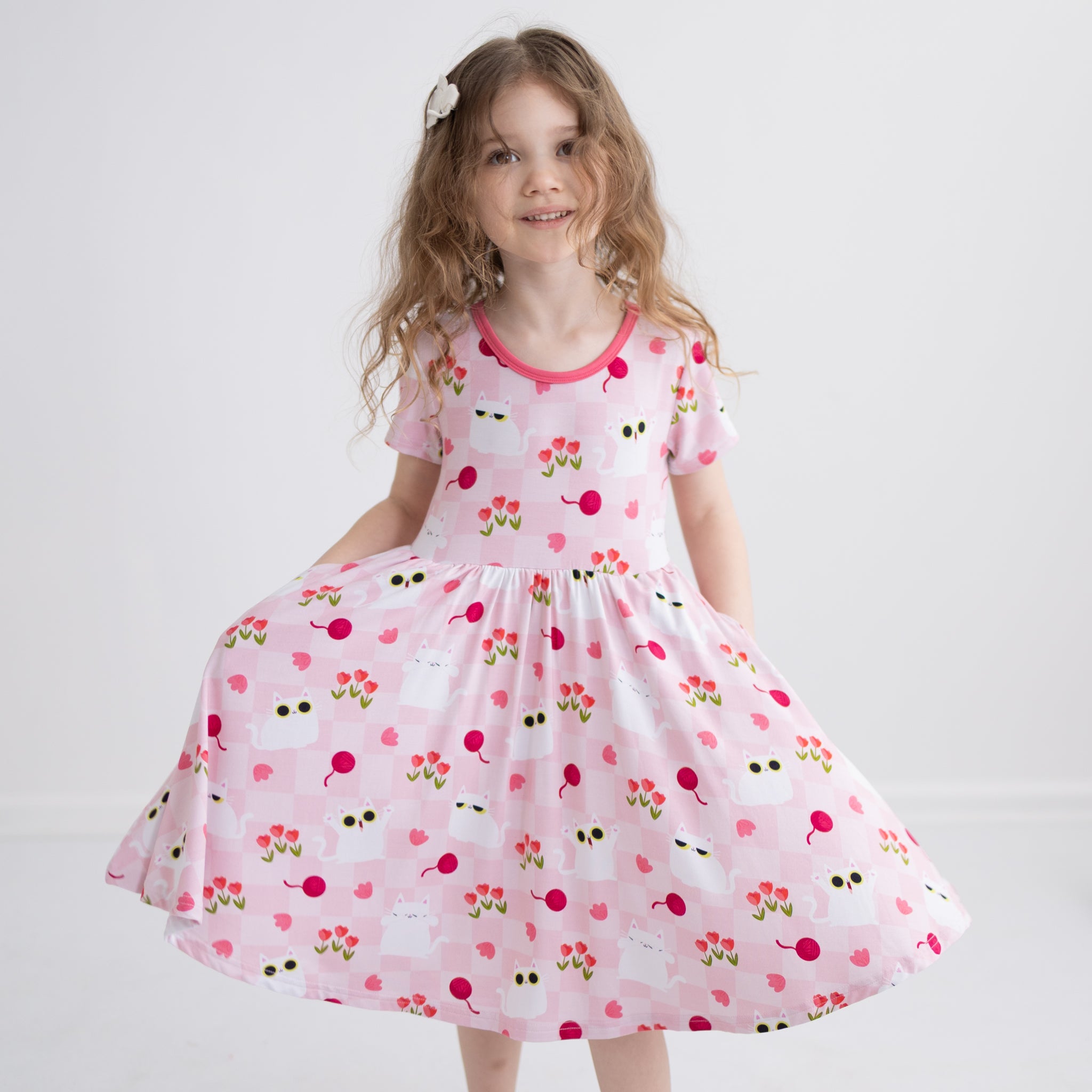 A girl wears a pink short-sleeved dress with a white cat and tulip pattern and side pockets.