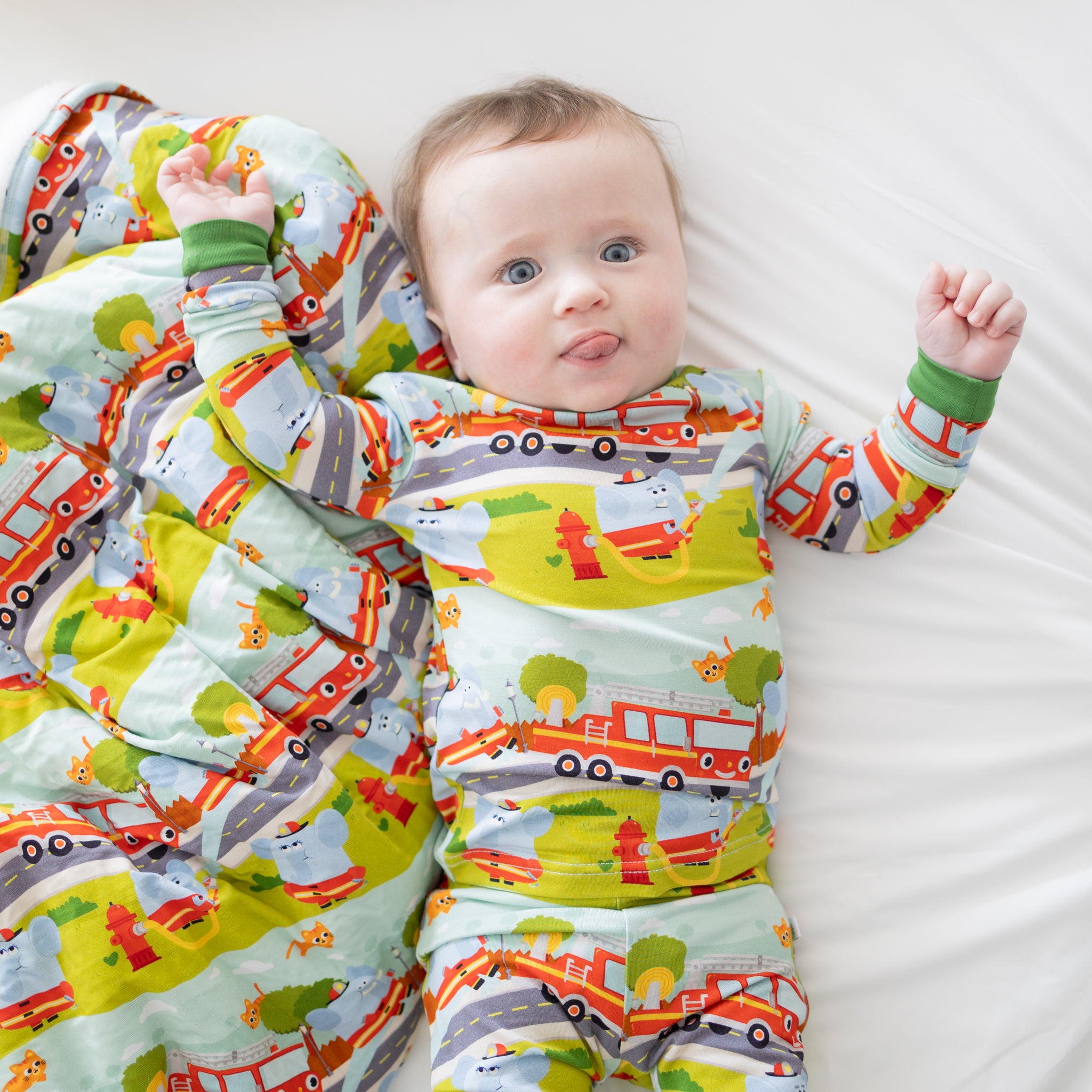 A baby lies on a soft blanket and matching pajamas featuring a colorful firetruck pattern.