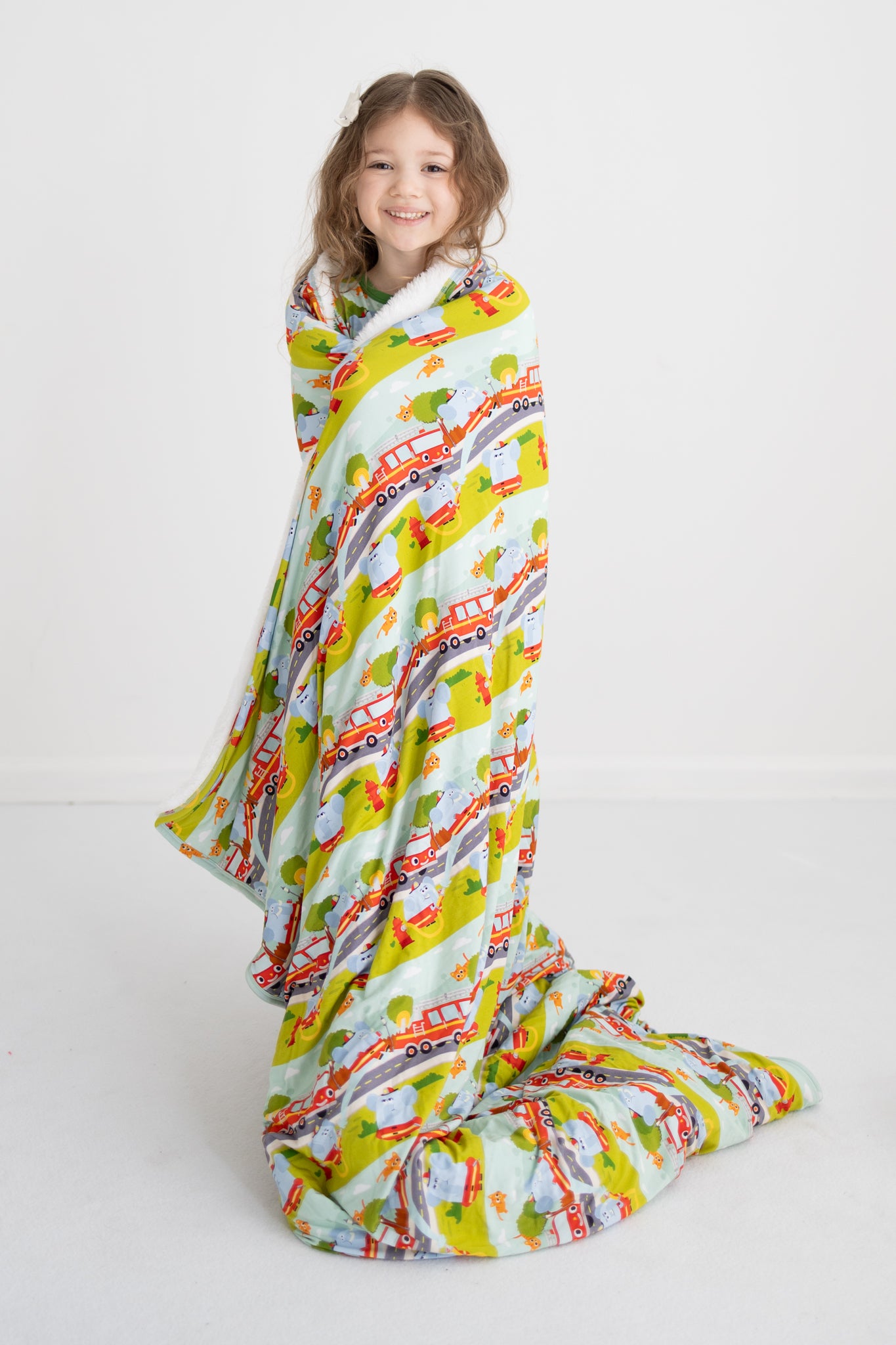 A young girl wrapped in a soft blanket featuring a colorful firetruck and character print.