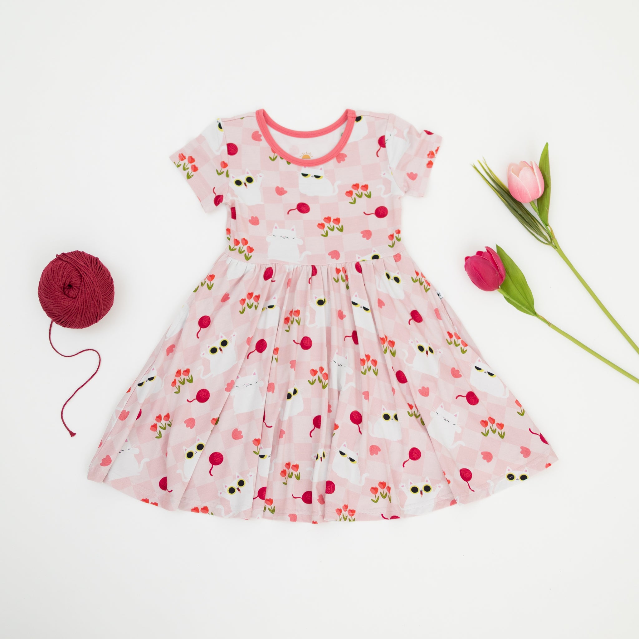A pink short-sleeved dress with a white cat and tulip pattern, displayed with yarn and tulips.