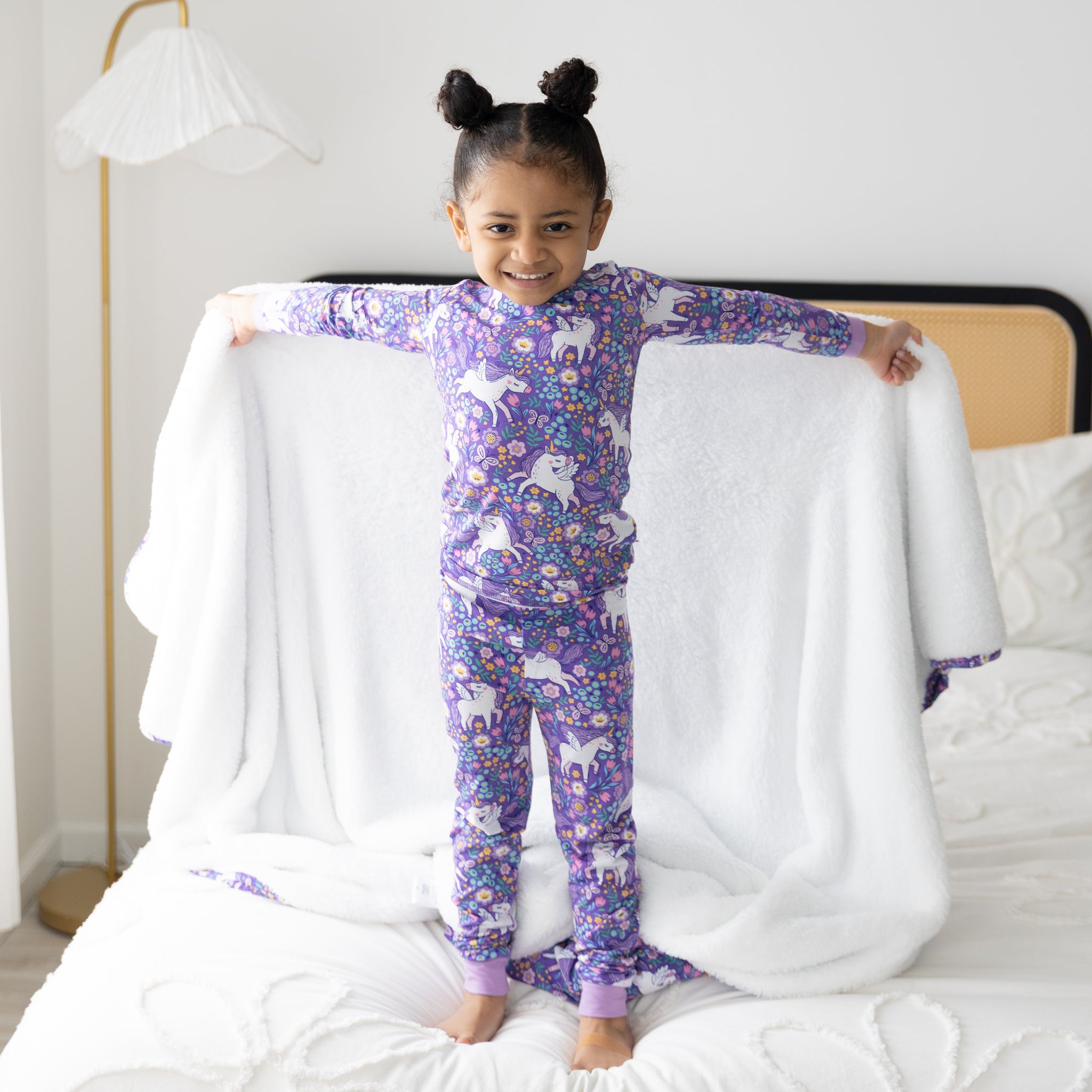 A young girl stands on a bed wearing a purple long-sleeve pajama set featuring a unicorn print.