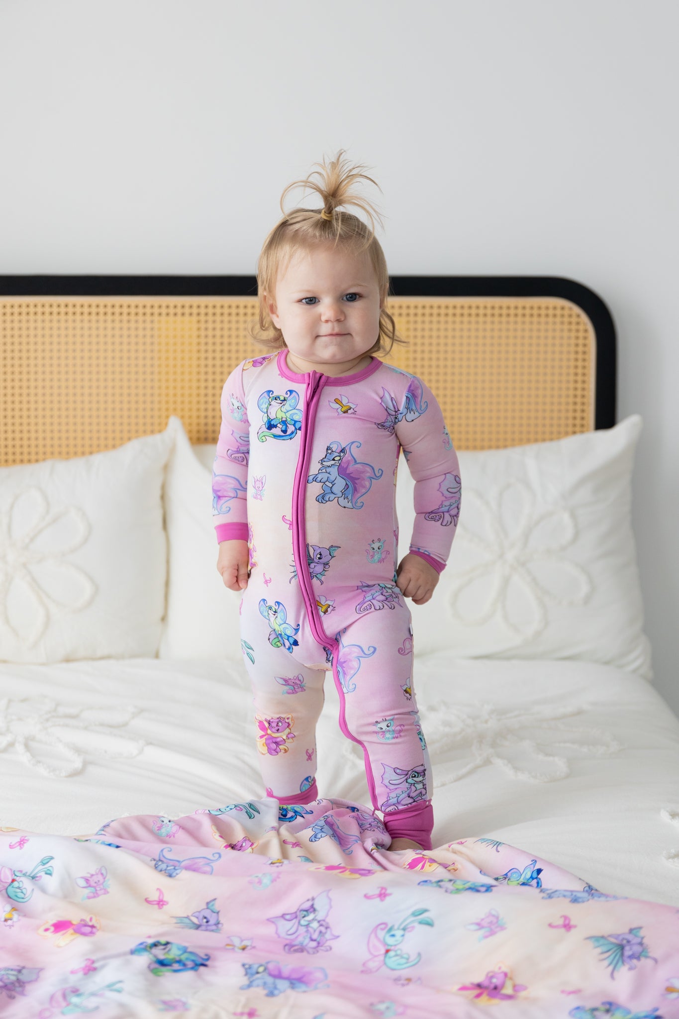 A toddler stands on a bed wearing a pink long-sleeve zip-up pajama featuring a Neopets Faerie print.