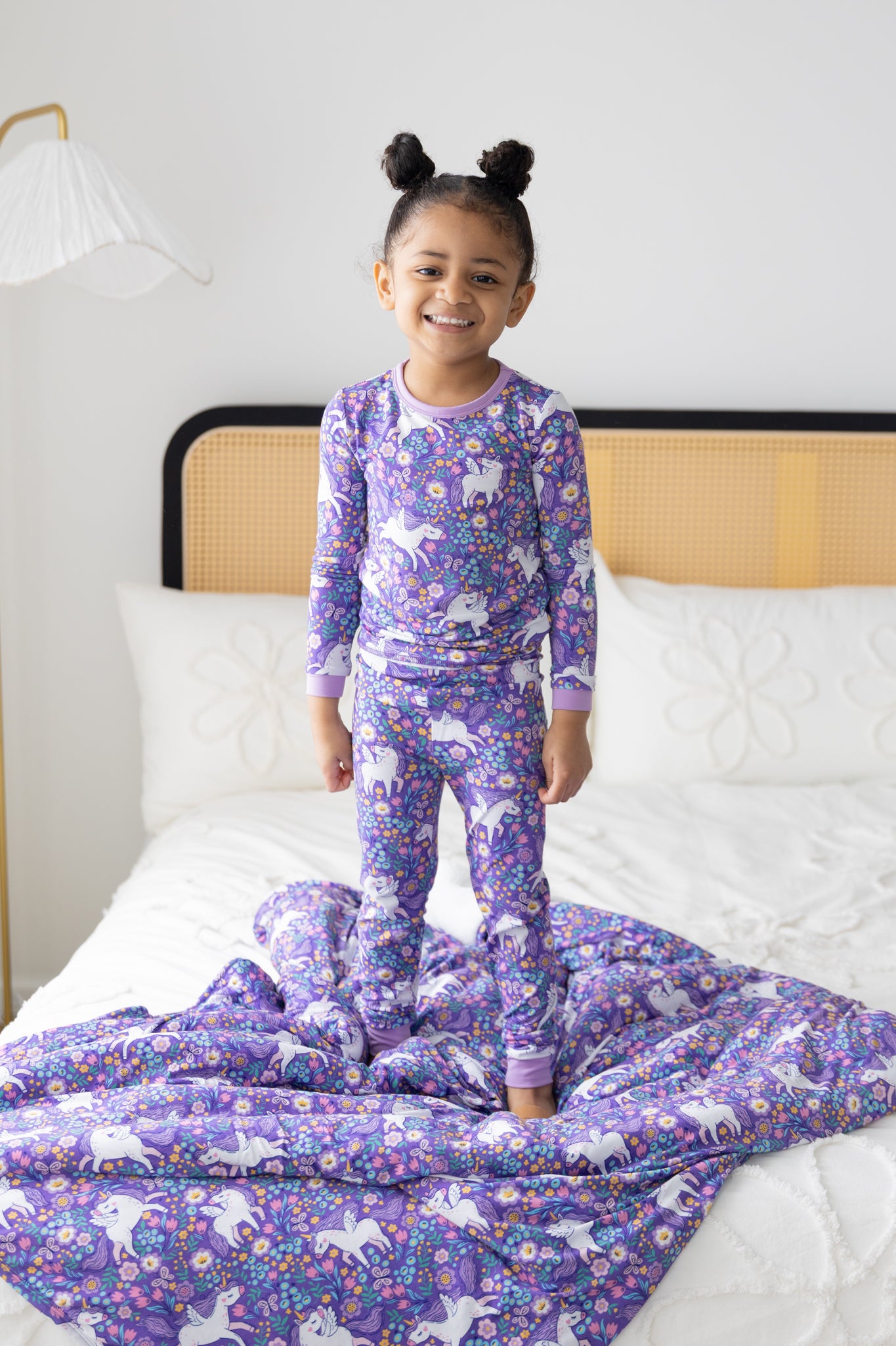 A young girl stands on a bed wearing a purple long-sleeve pajama set featuring a unicorn print.