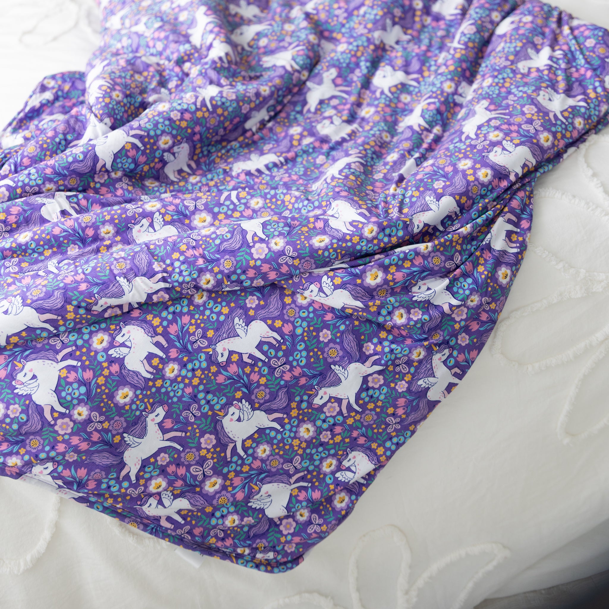A purple plush blanket featuring a repeating pattern of white winged unicorns and colorful flowers.