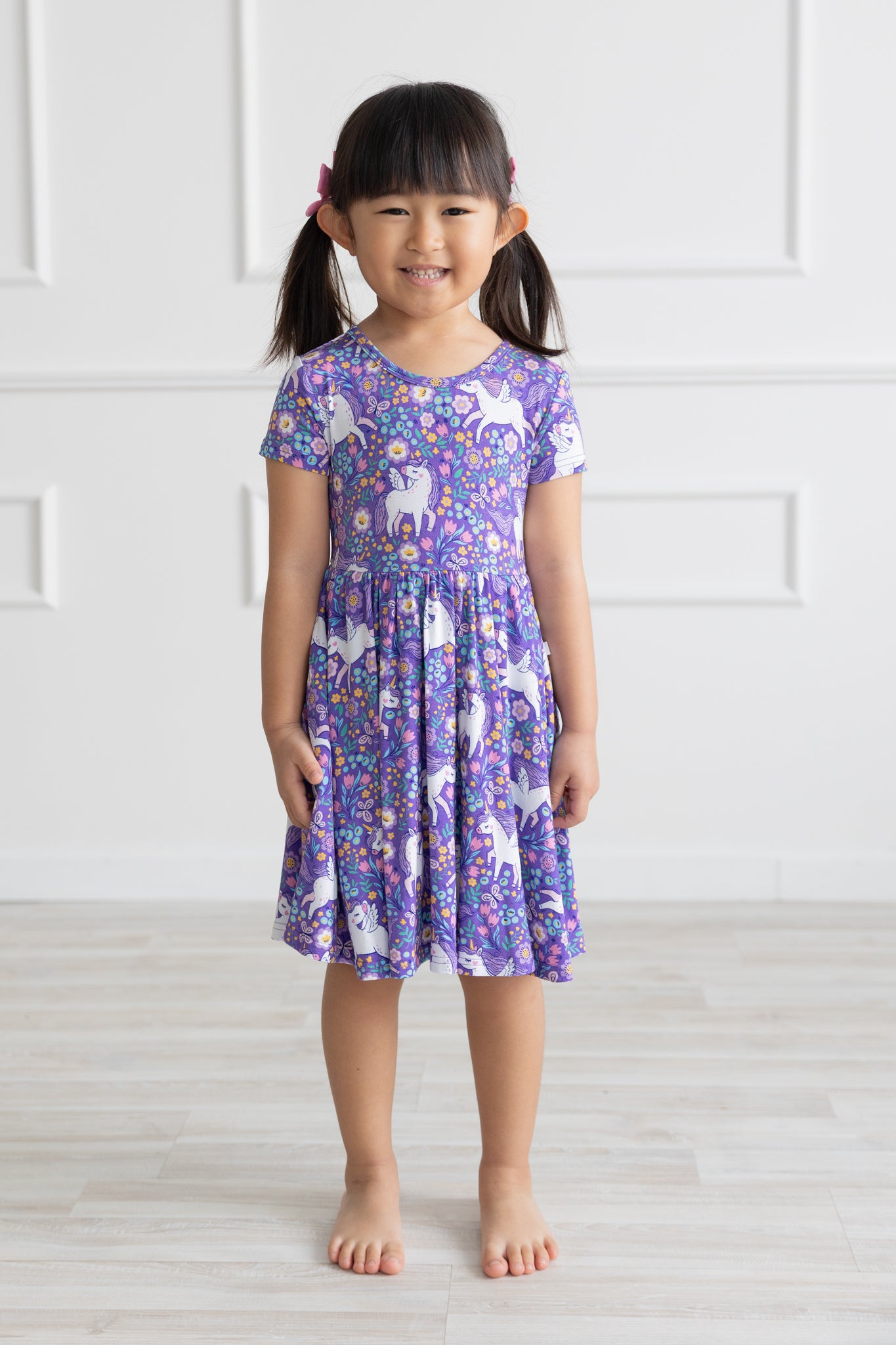 A young girl wears a purple short-sleeve dress featuring a white unicorn and floral print.