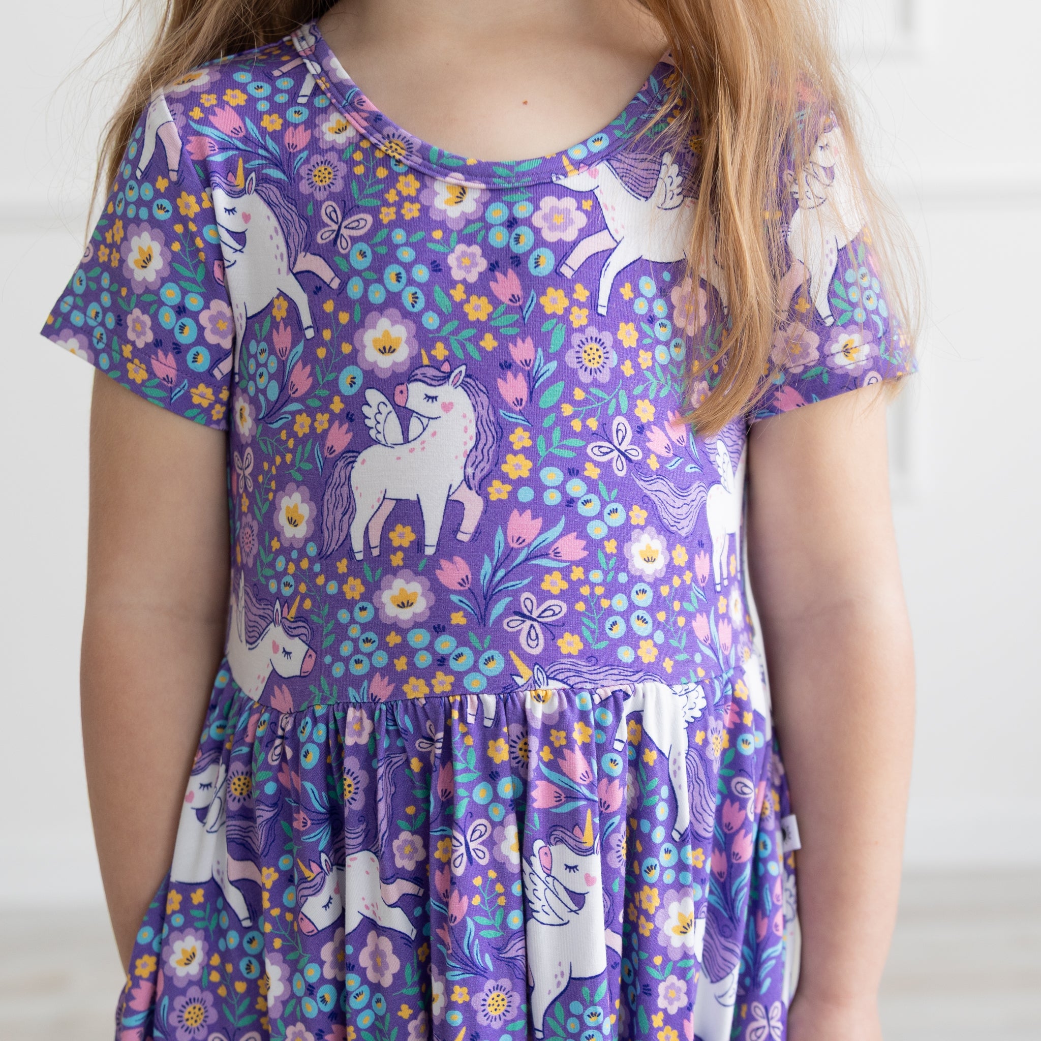 A child wears a purple short-sleeved dress featuring a repeating pattern of white unicorns and colorful flowers.