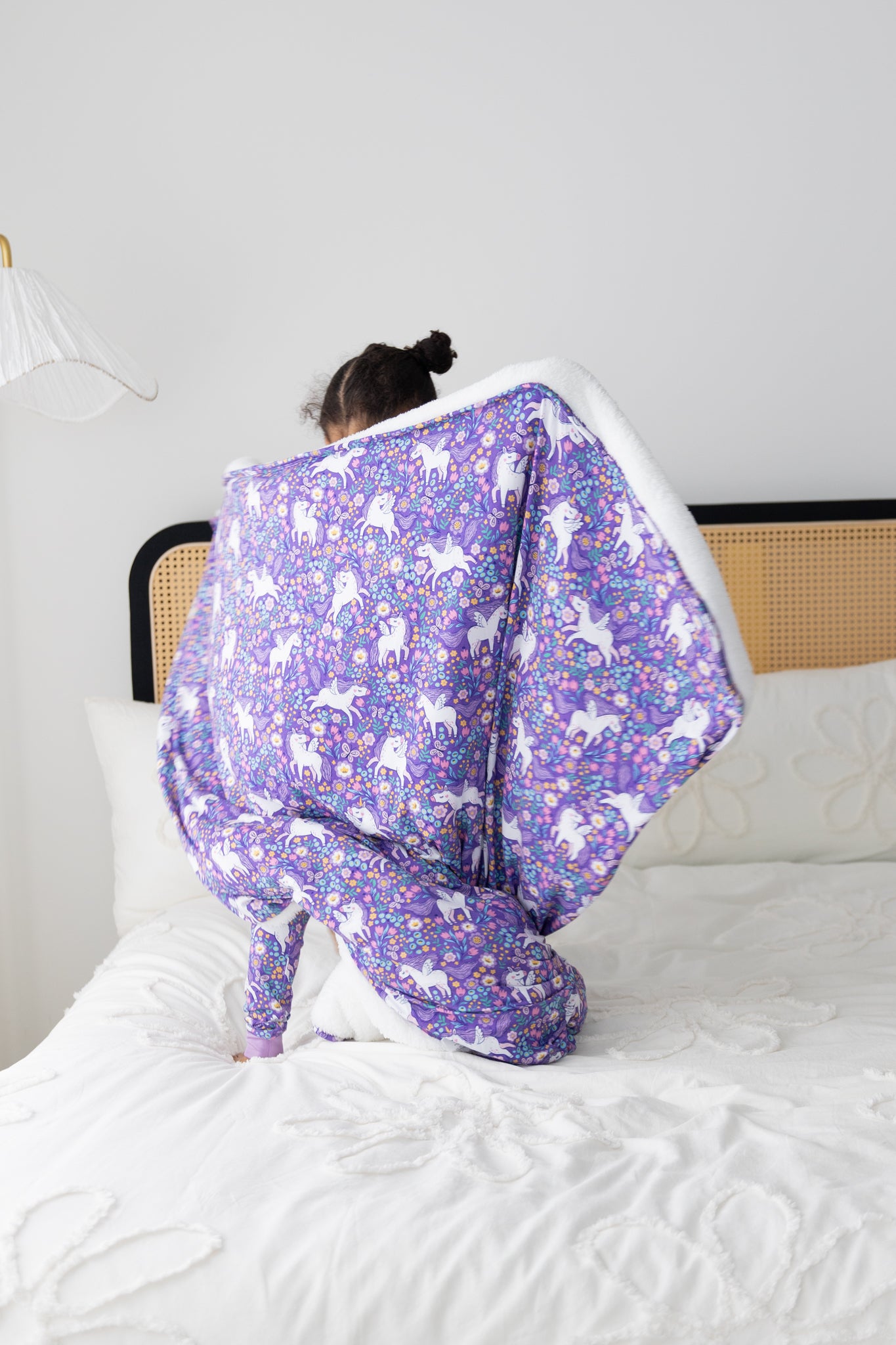 A child sits on a bed holding a purple unicorn-patterned plush blanket with a white fleece lining.