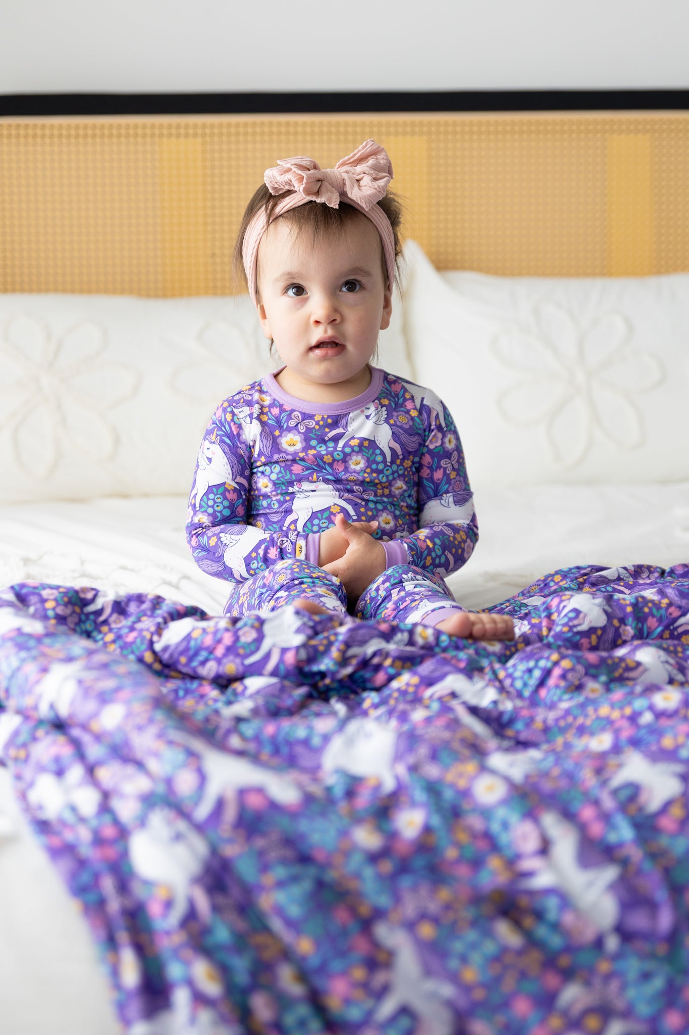 A baby sits on a bed wearing a purple long-sleeve pajama set featuring a unicorn and floral print.
