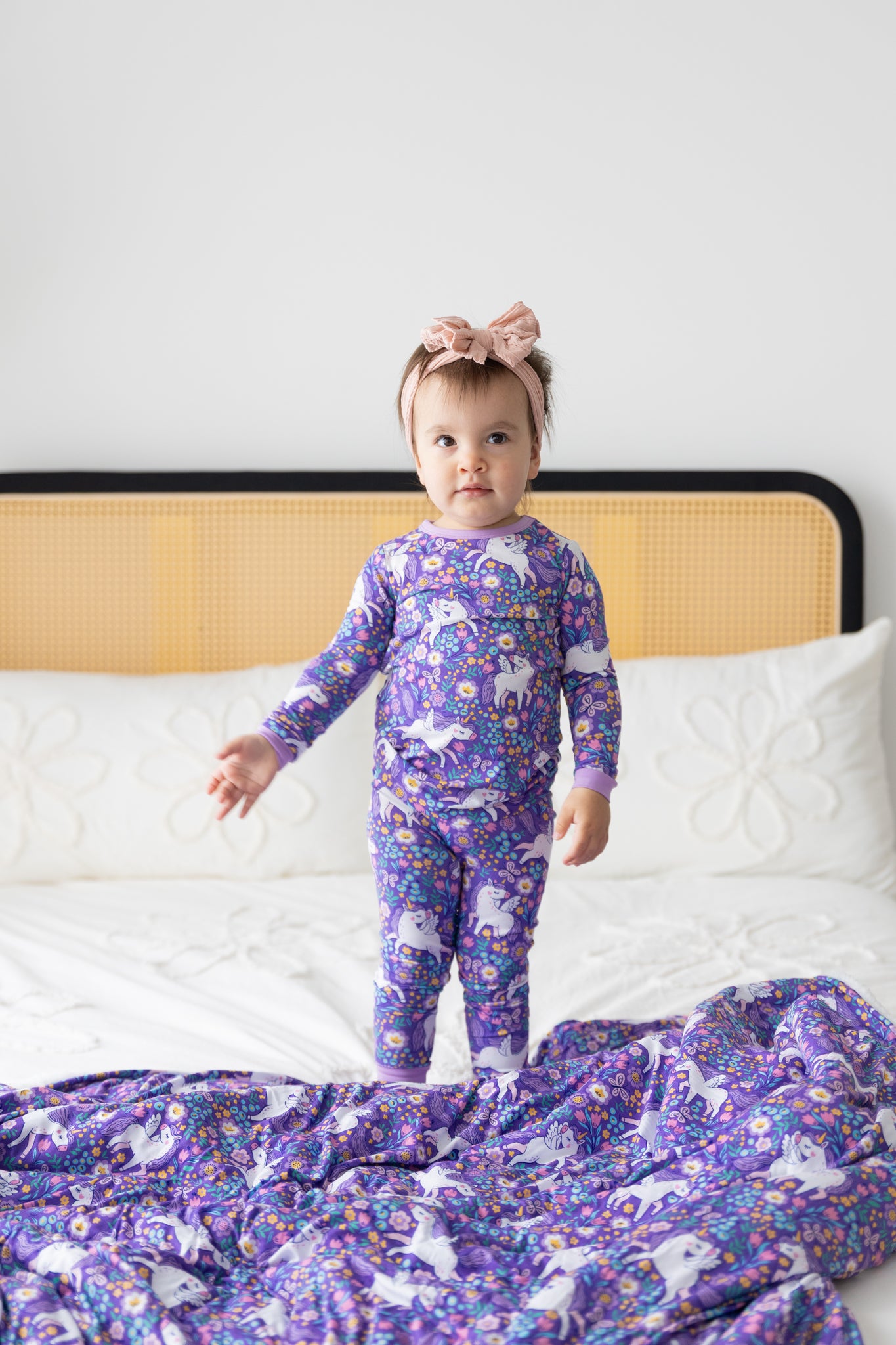 A toddler stands on a bed wearing a purple long-sleeve pajama set featuring a unicorn floral print.