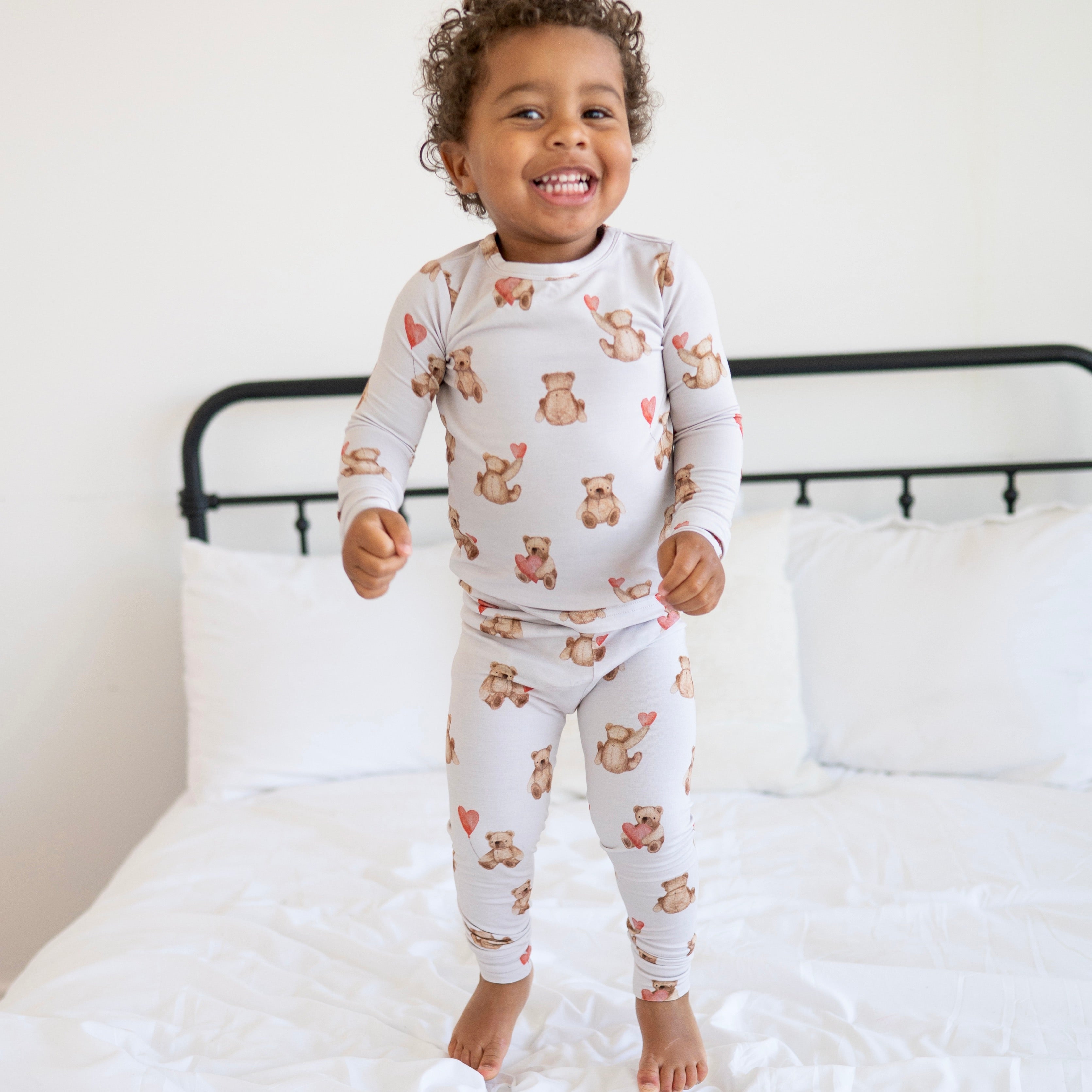 A young child smiles while wearing a light grey pajama set with a teddy bear and heart pattern.