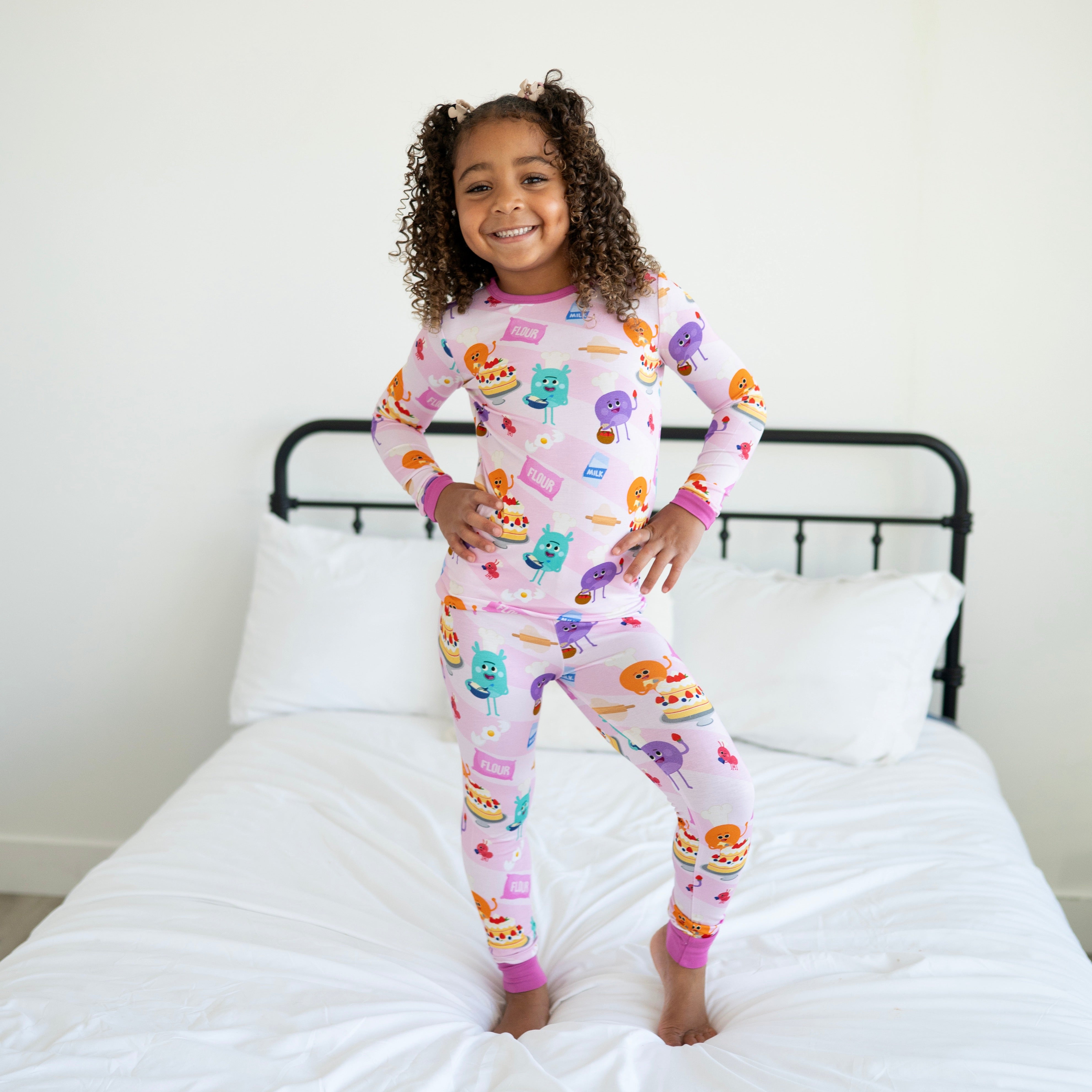 A young girl smiles on a bed wearing pink pajamas with a baking-themed print.