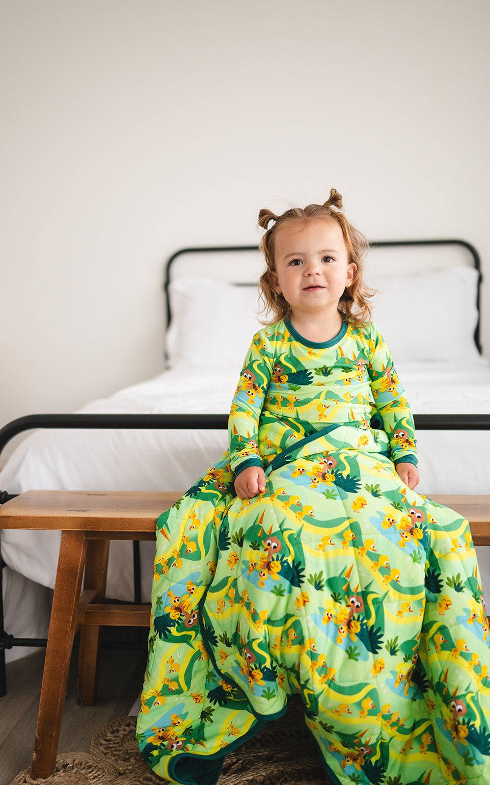 A toddler sits on a bench wearing a green long-sleeved shirt and blanket with a duckling print.
