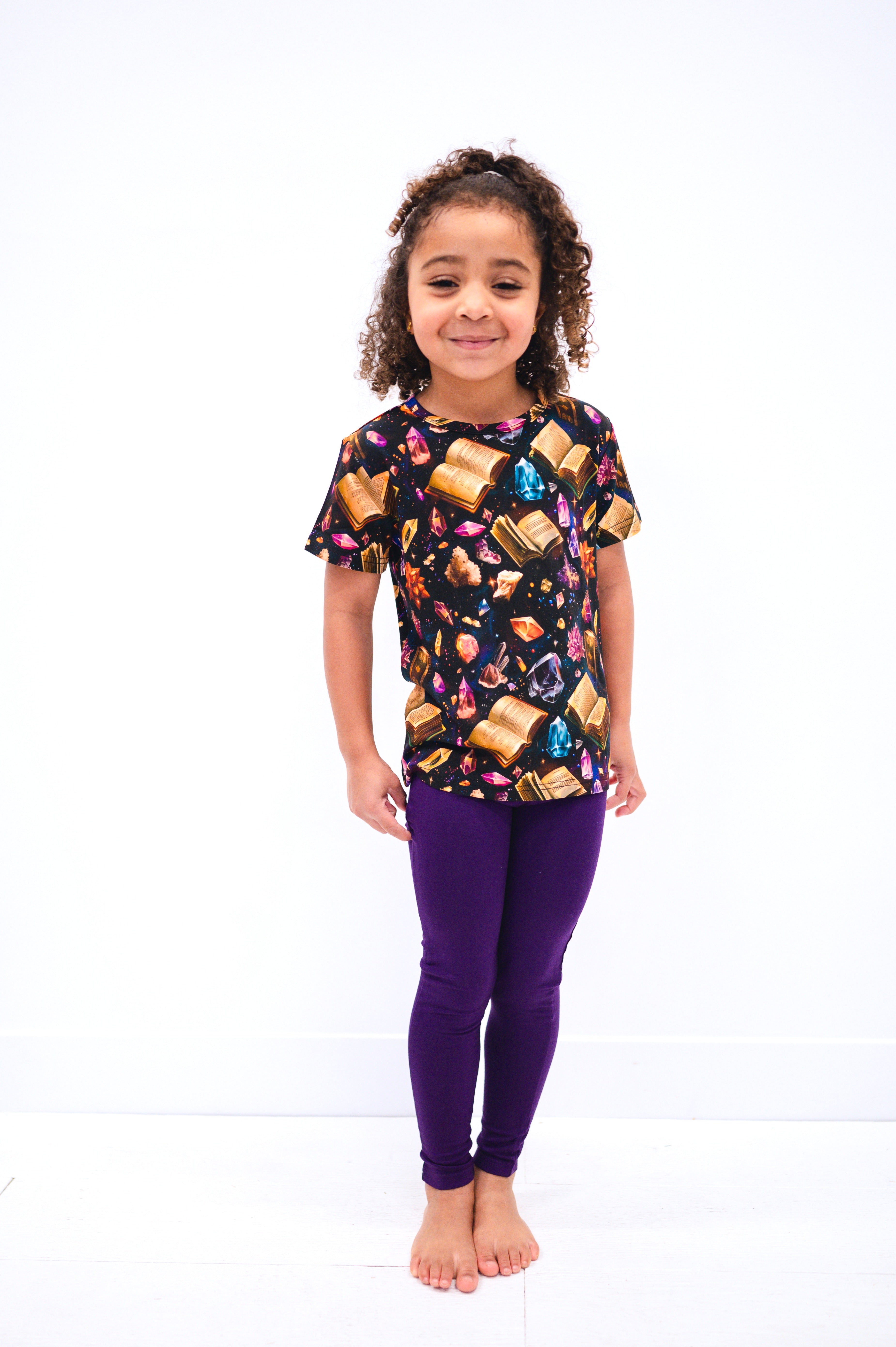 A young girl wears a black t-shirt with a book and crystal print and purple leggings.