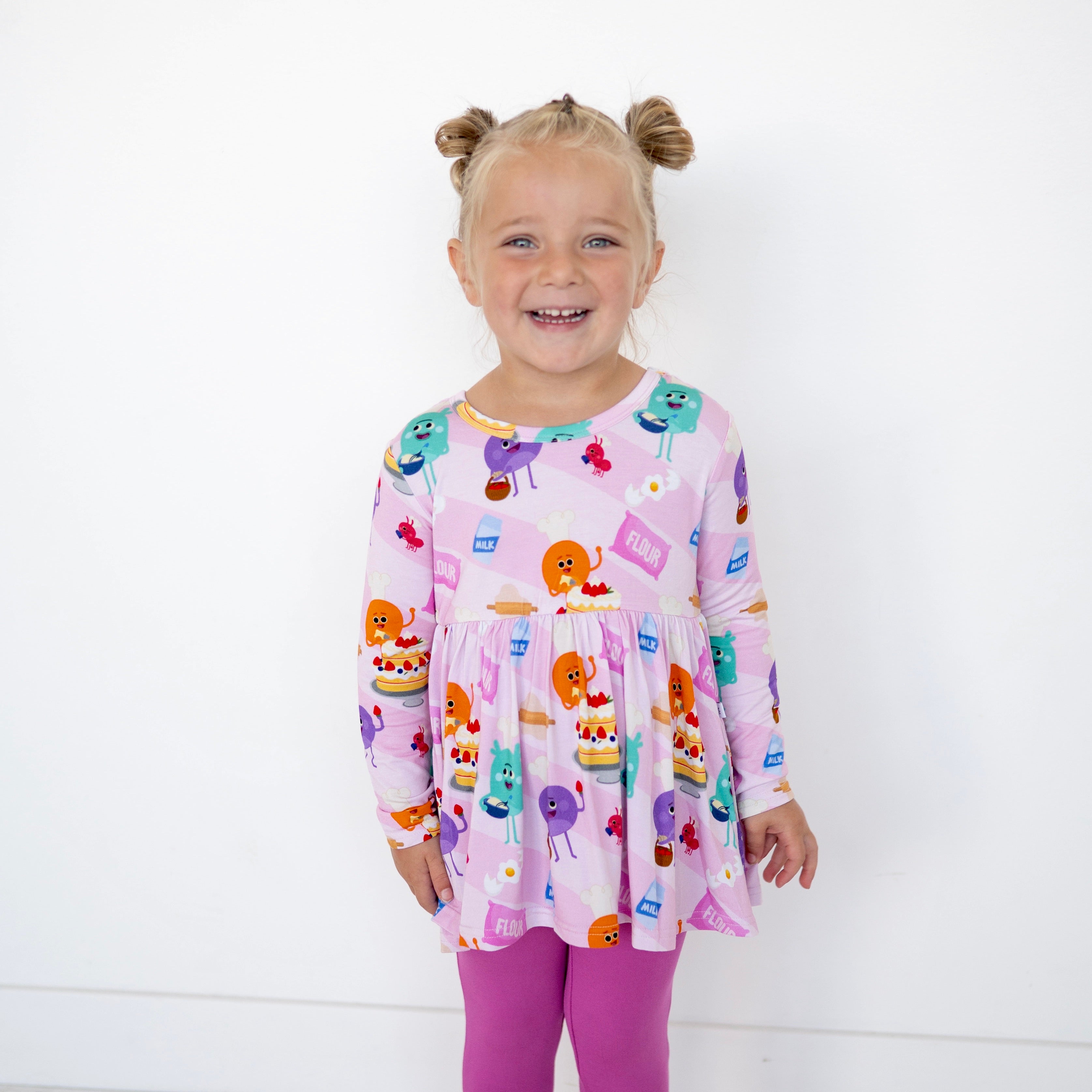 A young girl wears a pink long-sleeve peplum top with a baking-themed print and purple leggings.