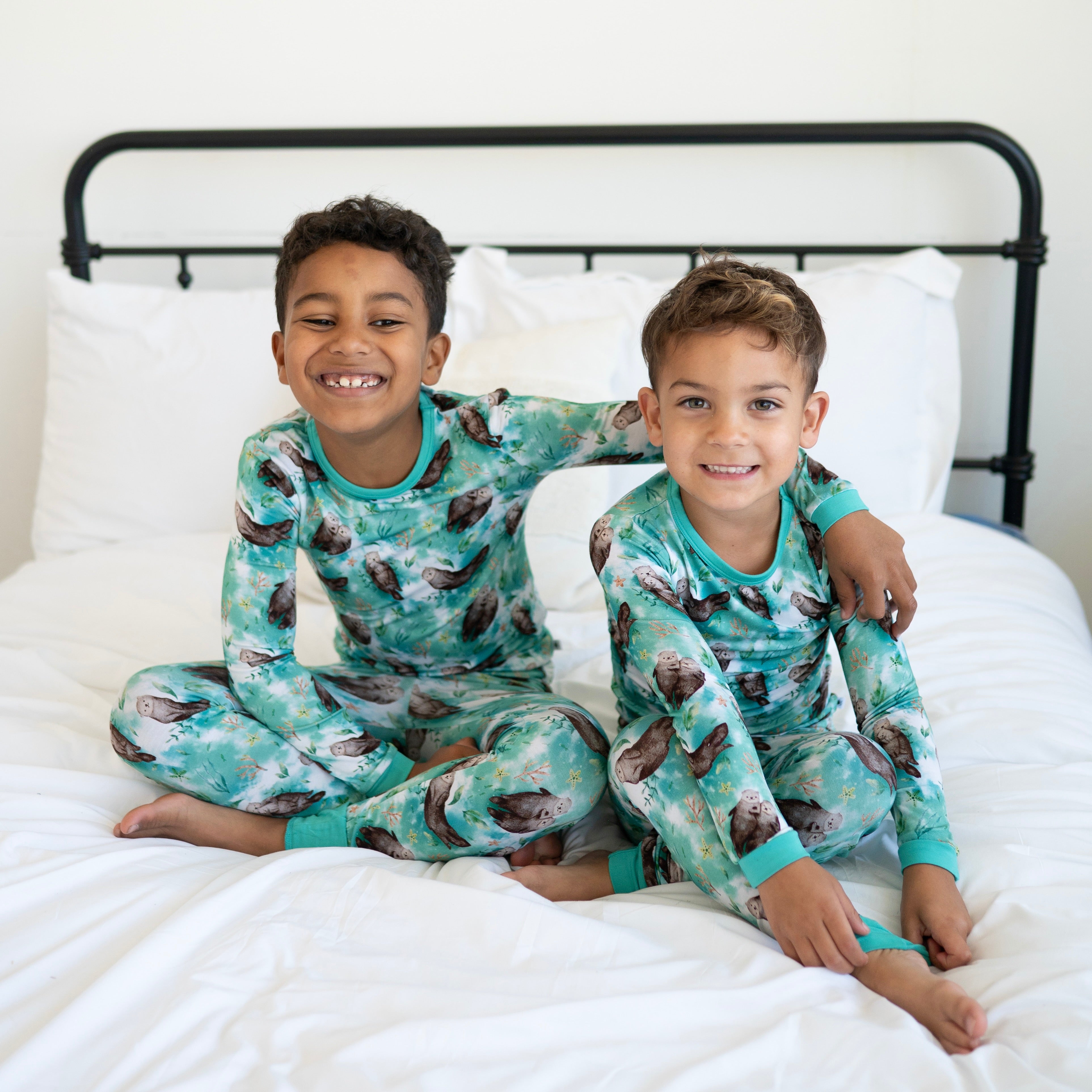 Two children sit on a bed wearing matching teal long-sleeve pajama sets with an otter print.