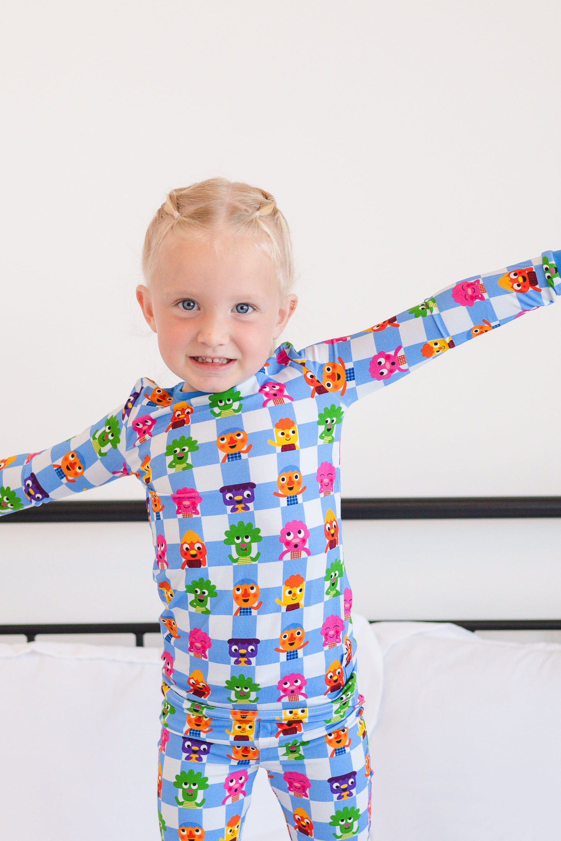A young girl in blue checkered pajamas with colorful monster characters stands on a bed.