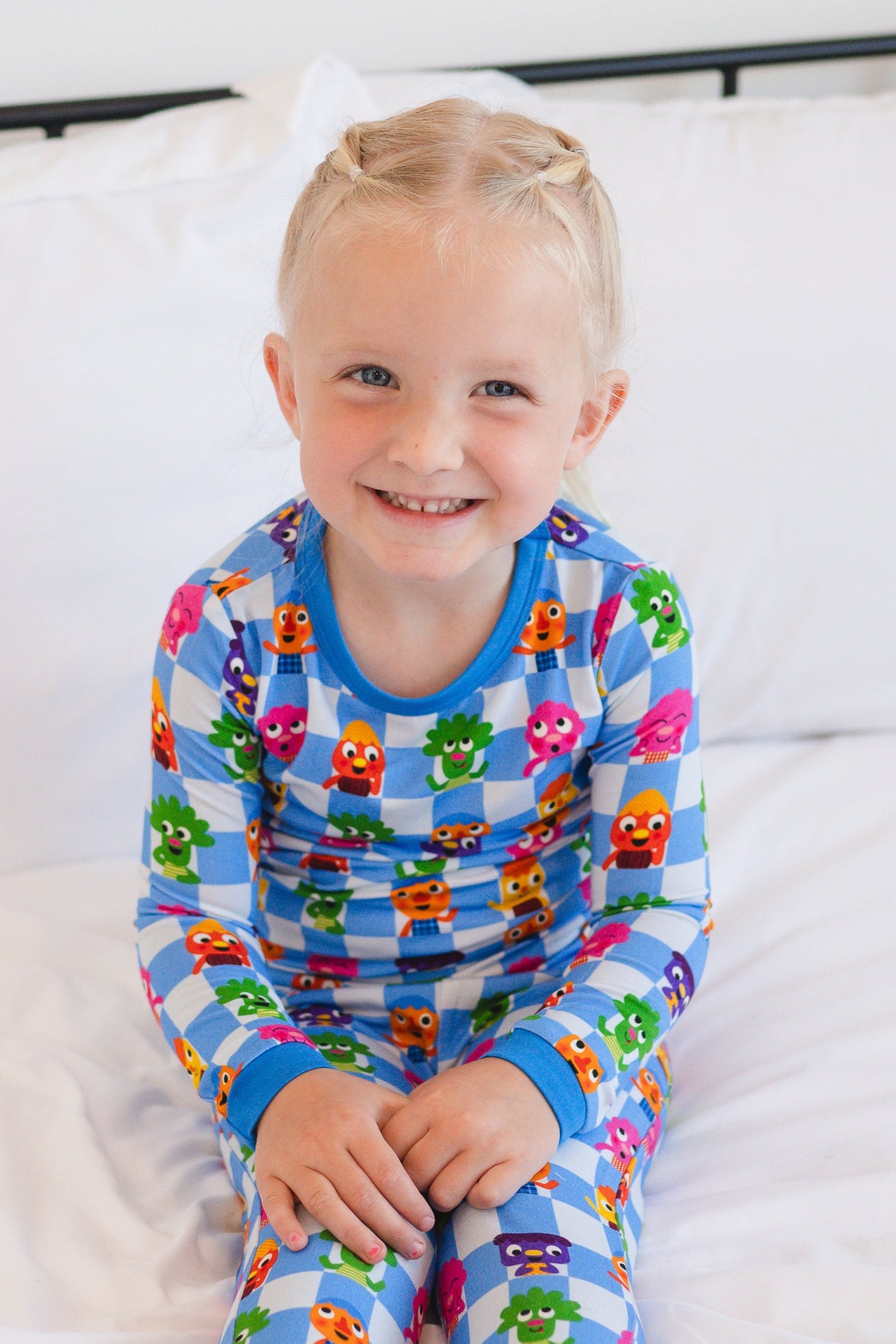 A smiling child sits on a white bed wearing blue and white checkered pajamas with colorful characters.