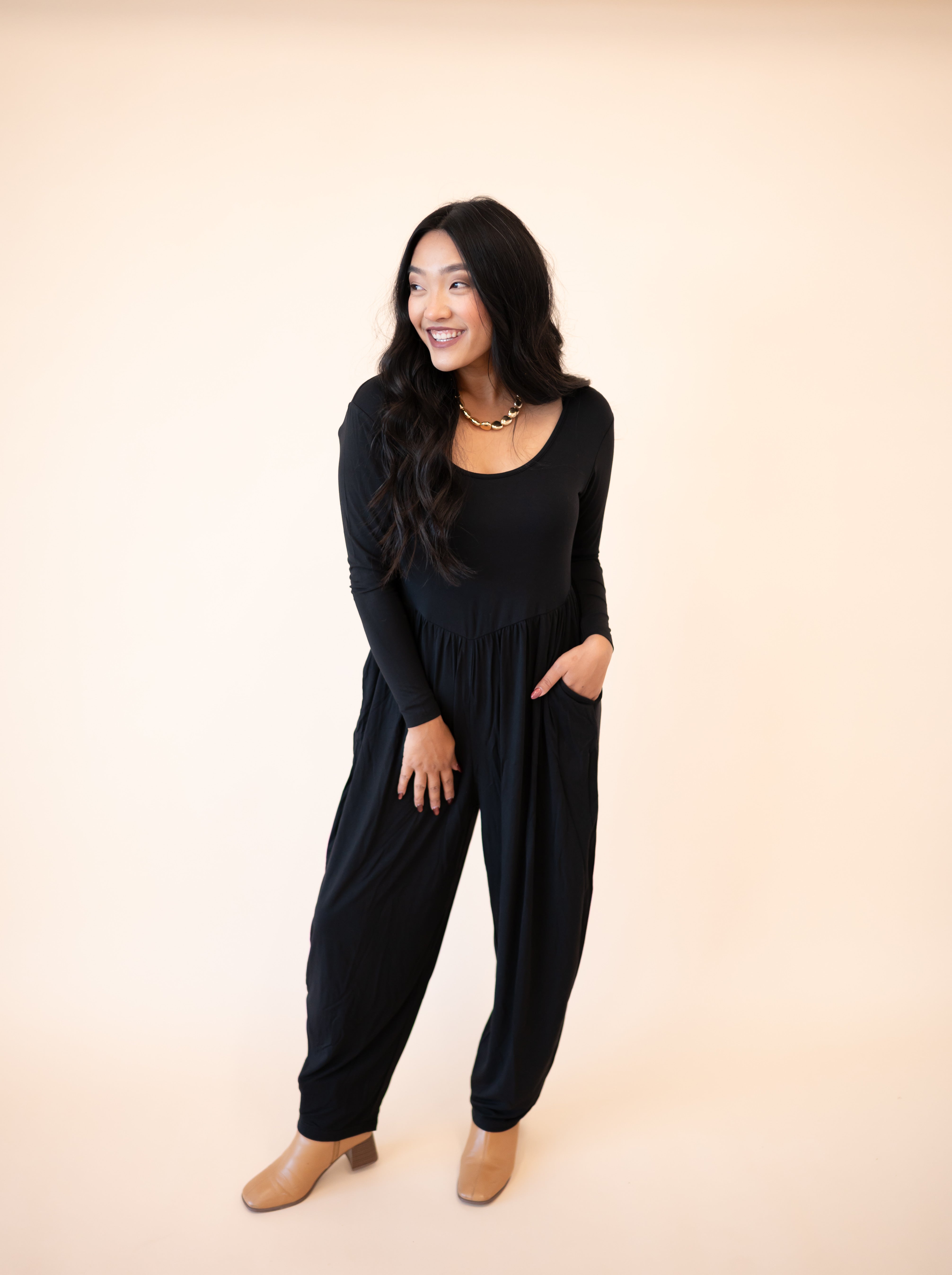 A woman smiles in a long-sleeve black Tencel Modal romper with a scoop neck and tapered legs.