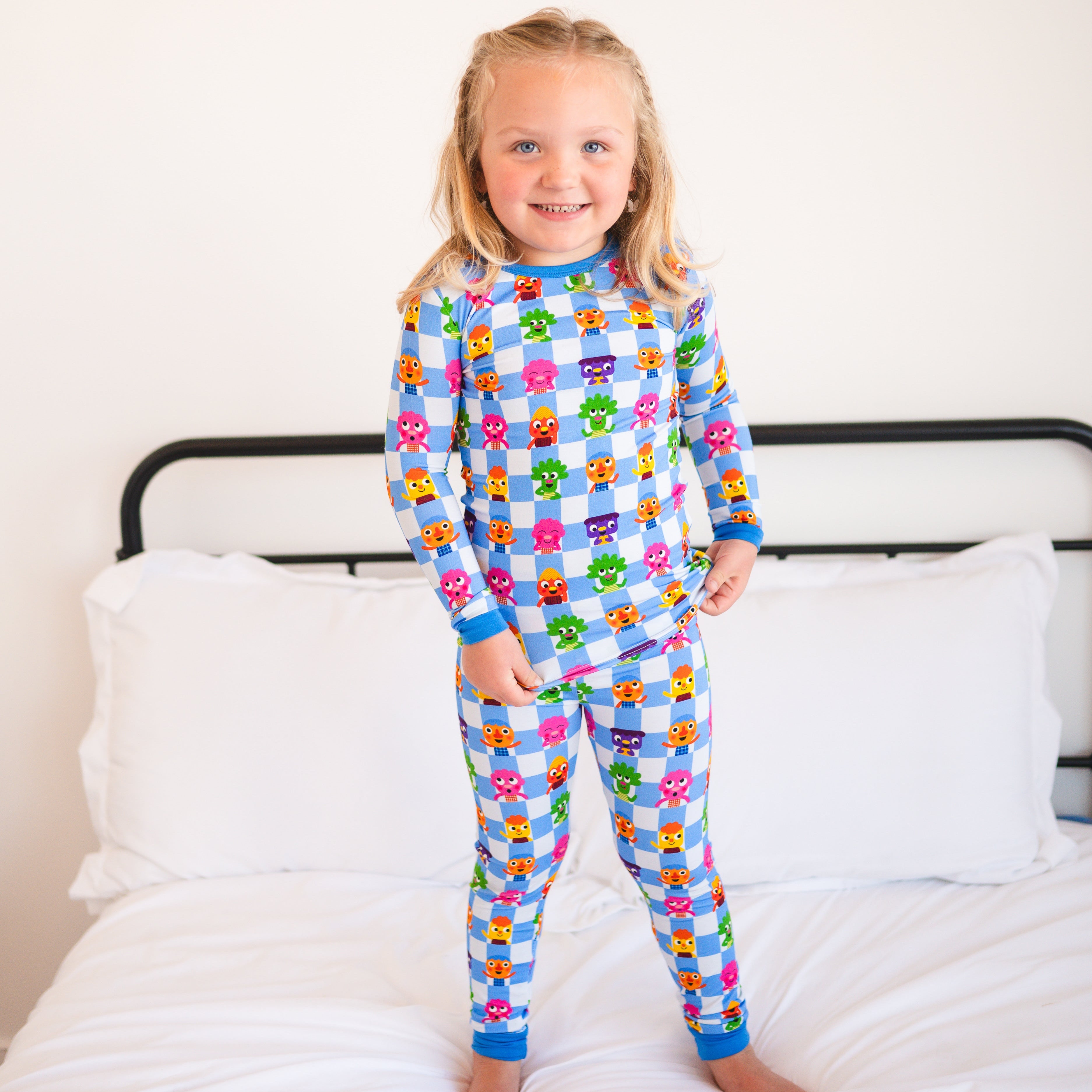 A young girl stands on a bed wearing blue and white checkered pajamas with cartoon characters.