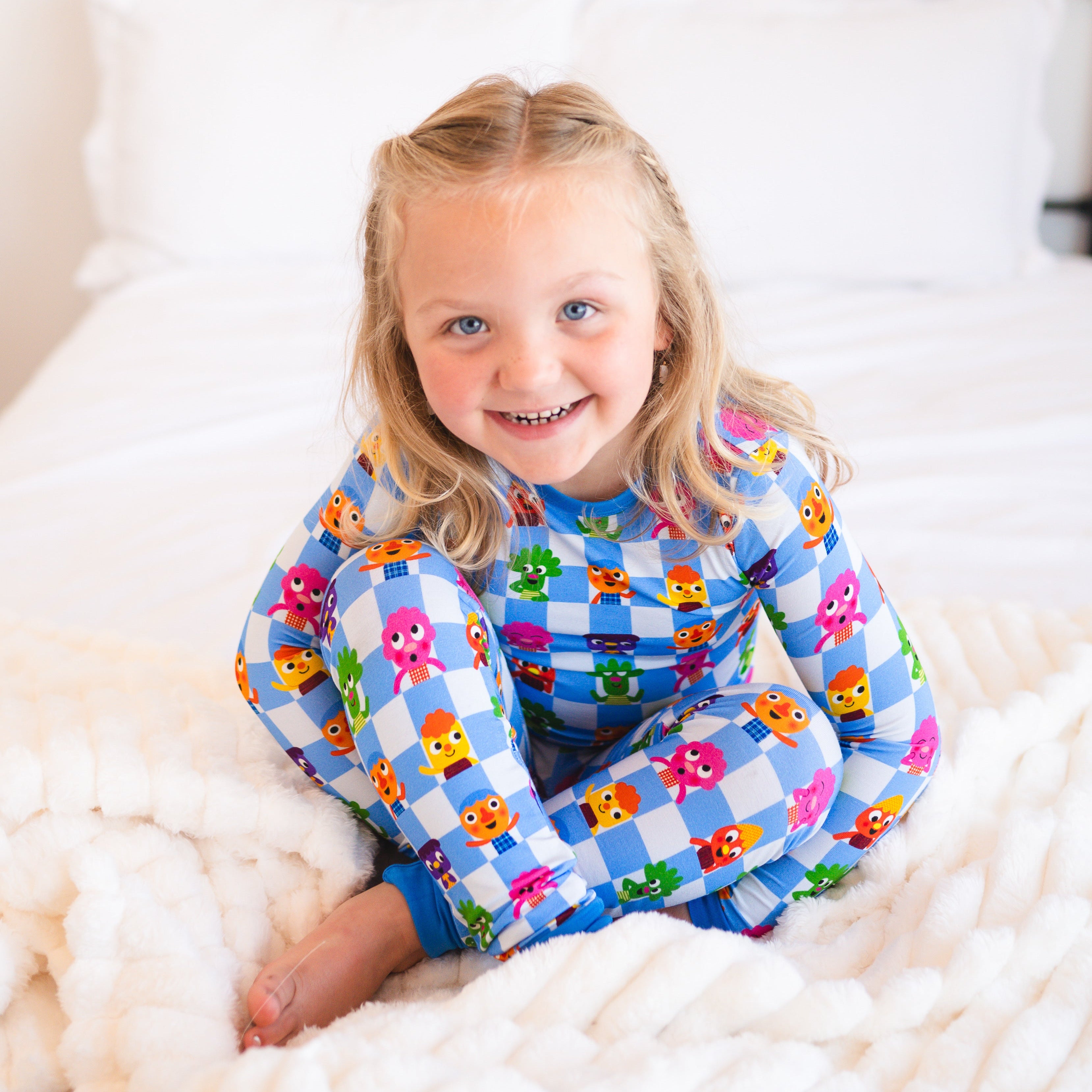 A young girl smiles while sitting on a fluffy blanket wearing blue checkered pajamas with cartoon characters.