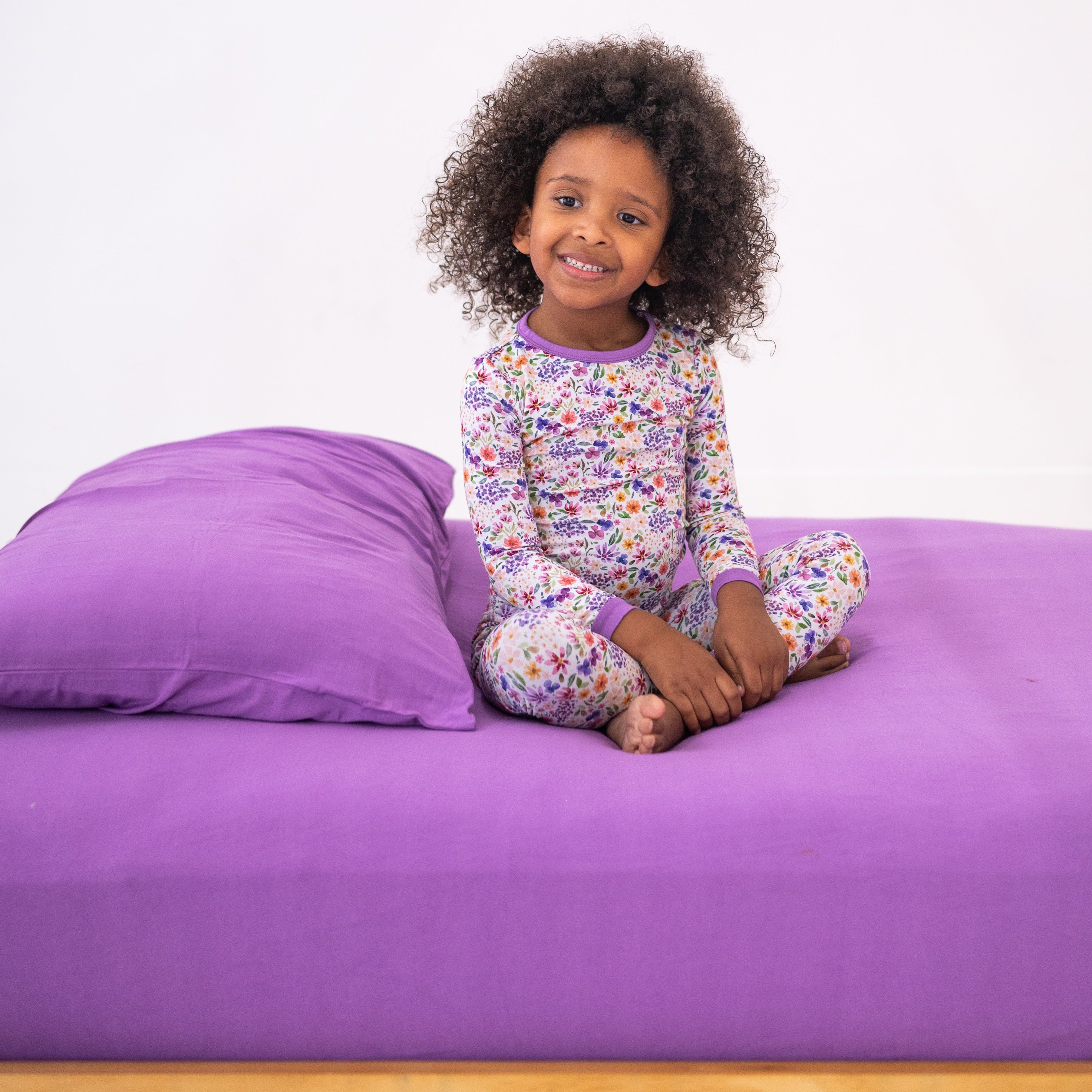 A young girl in floral pajamas sits on a lavender bed with a matching pillow.