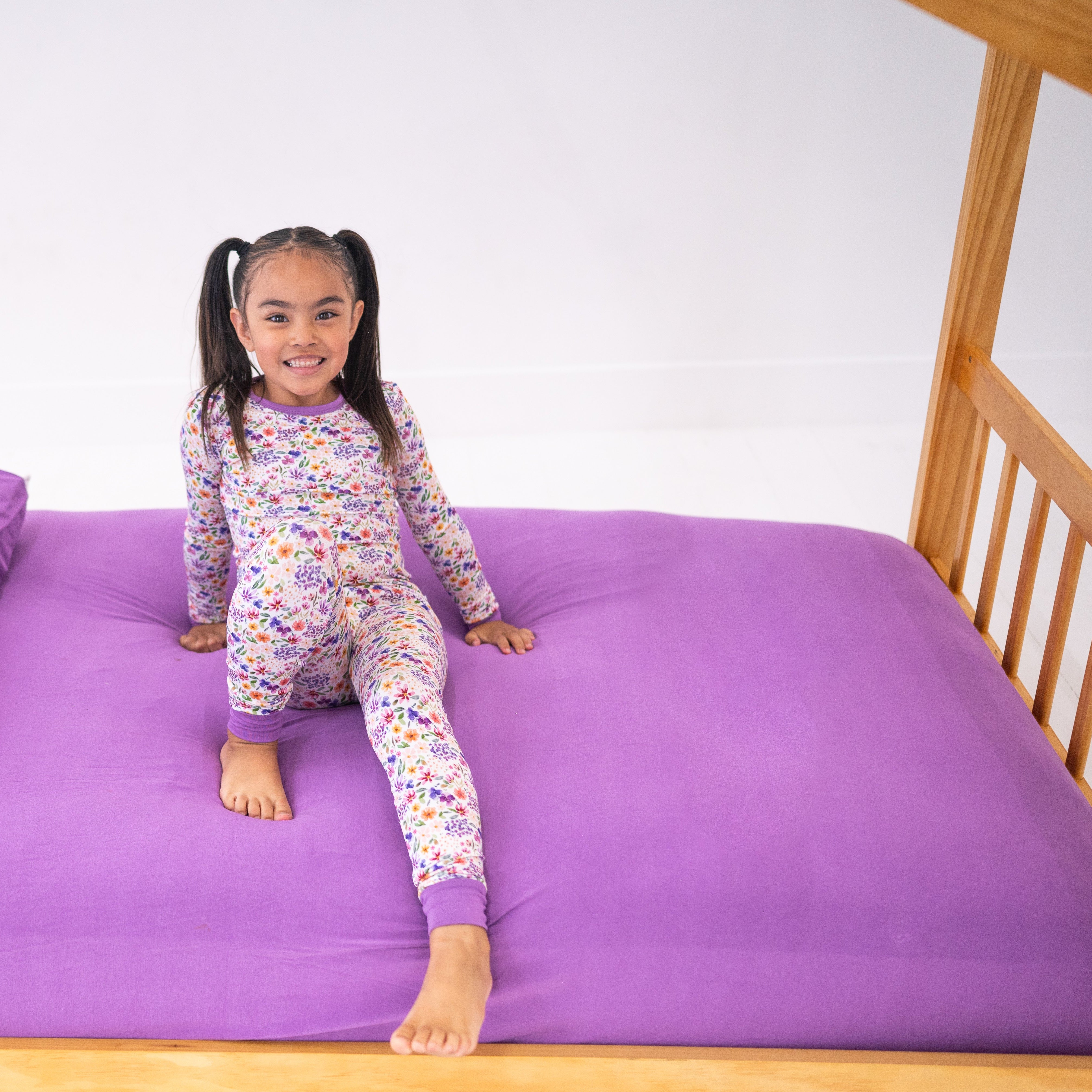 A young girl in floral pajamas sits on a purple fitted twin sheet.